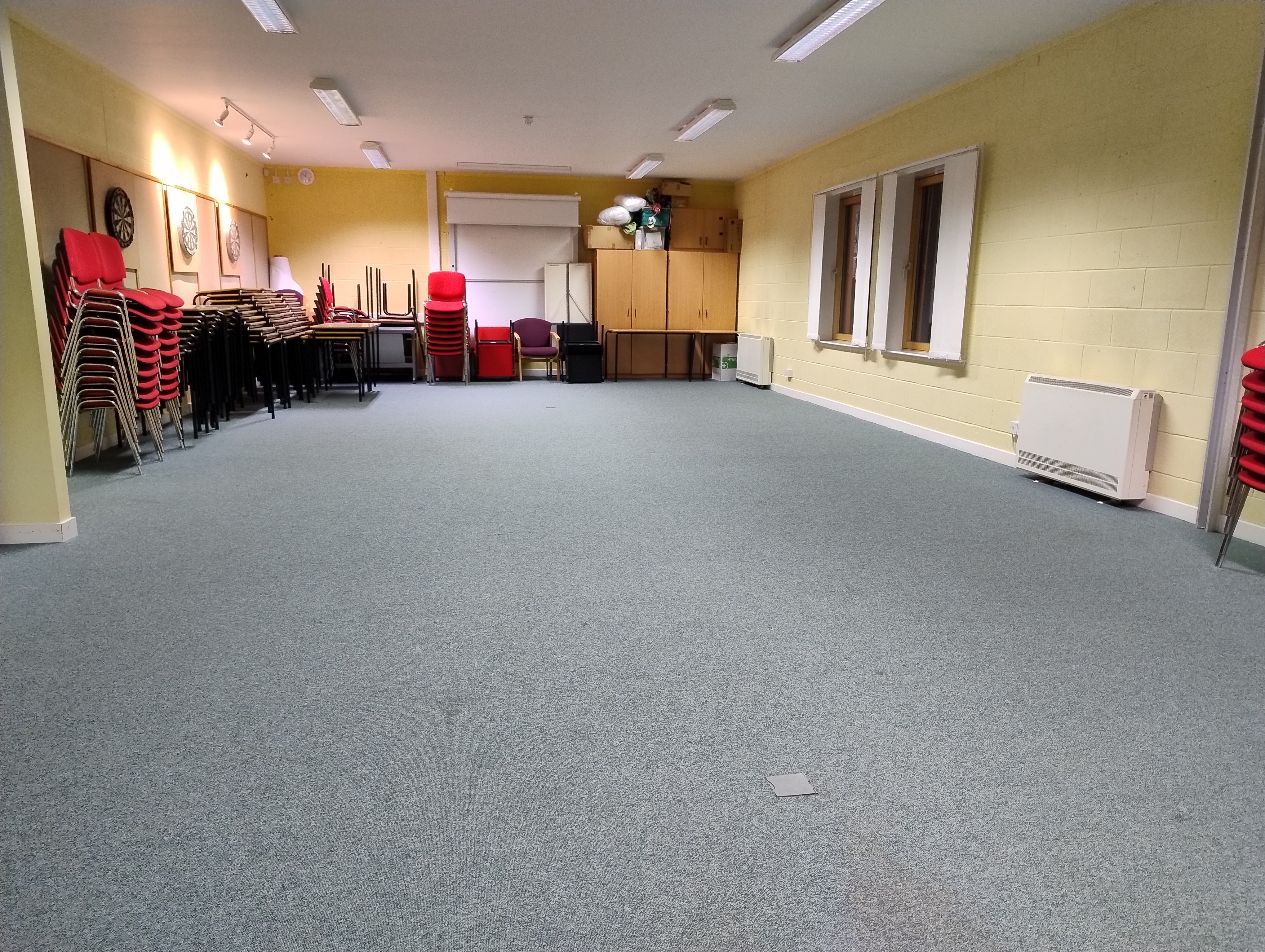 Spacious room featuring carpeted flooring, three dartboards mounted on the wall, and tables and chairs neatly stacked against the far wall.