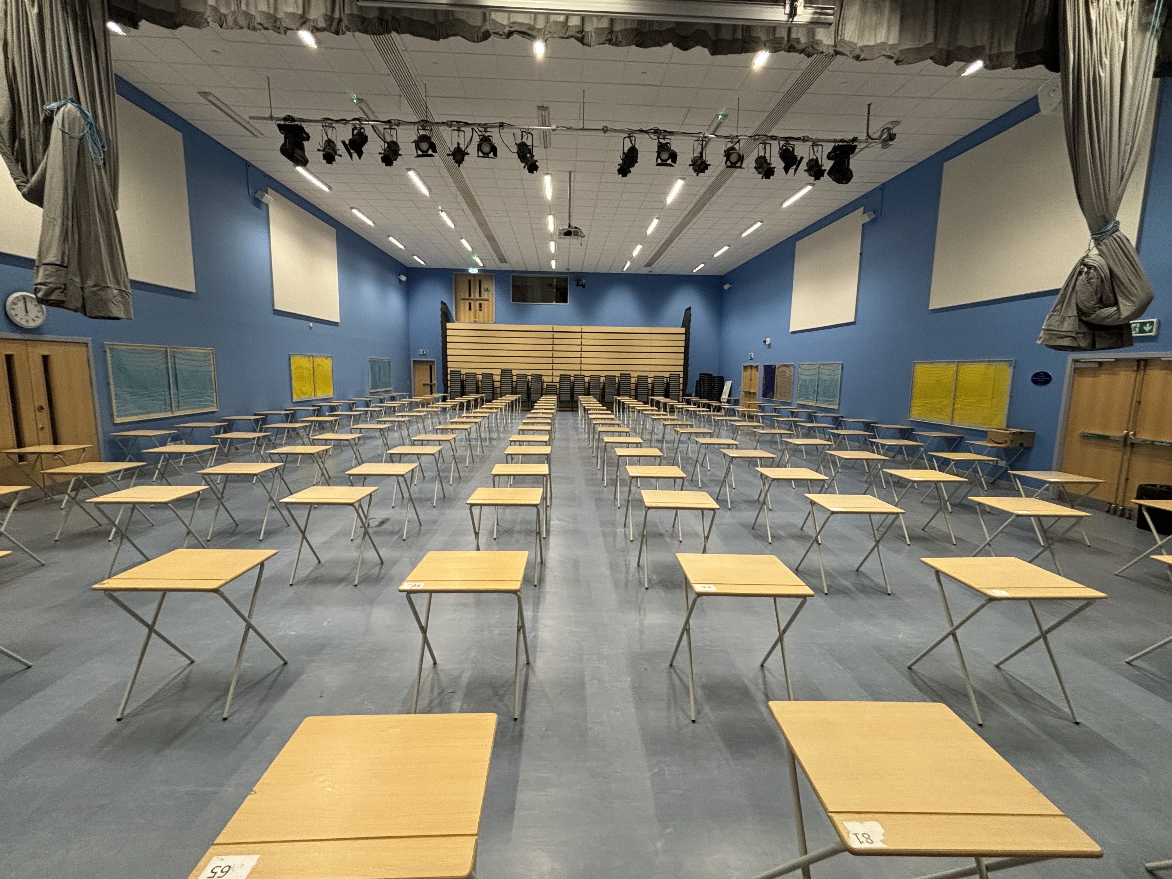A spacious hall features a ceiling-mounted lighting rig and whiteboards affixed to the side walls. The layout shows individual tables arranged for school examinations.