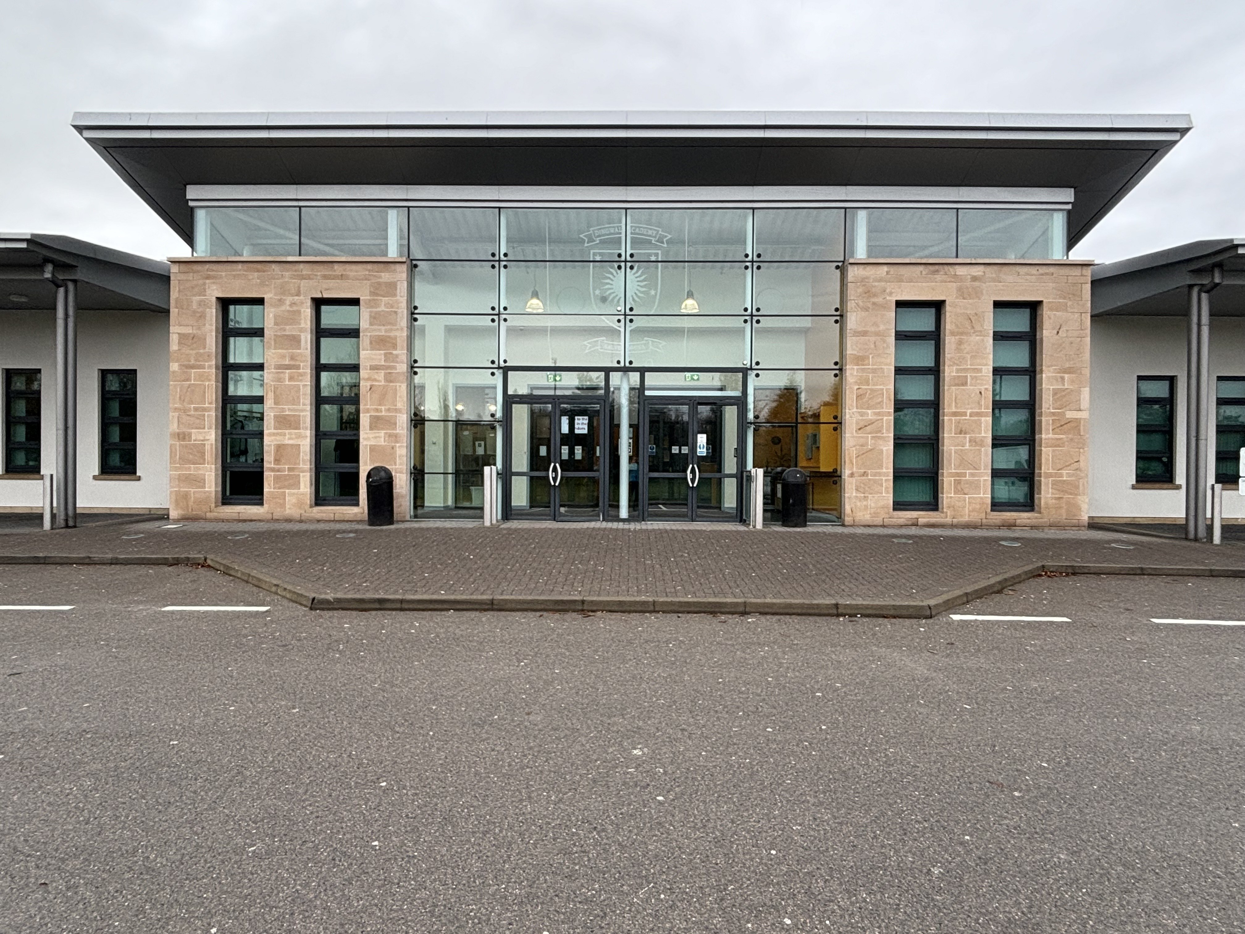 A modern building featuring a glass façade and entrance doors, with a curbed pavement leading up to the entrance.