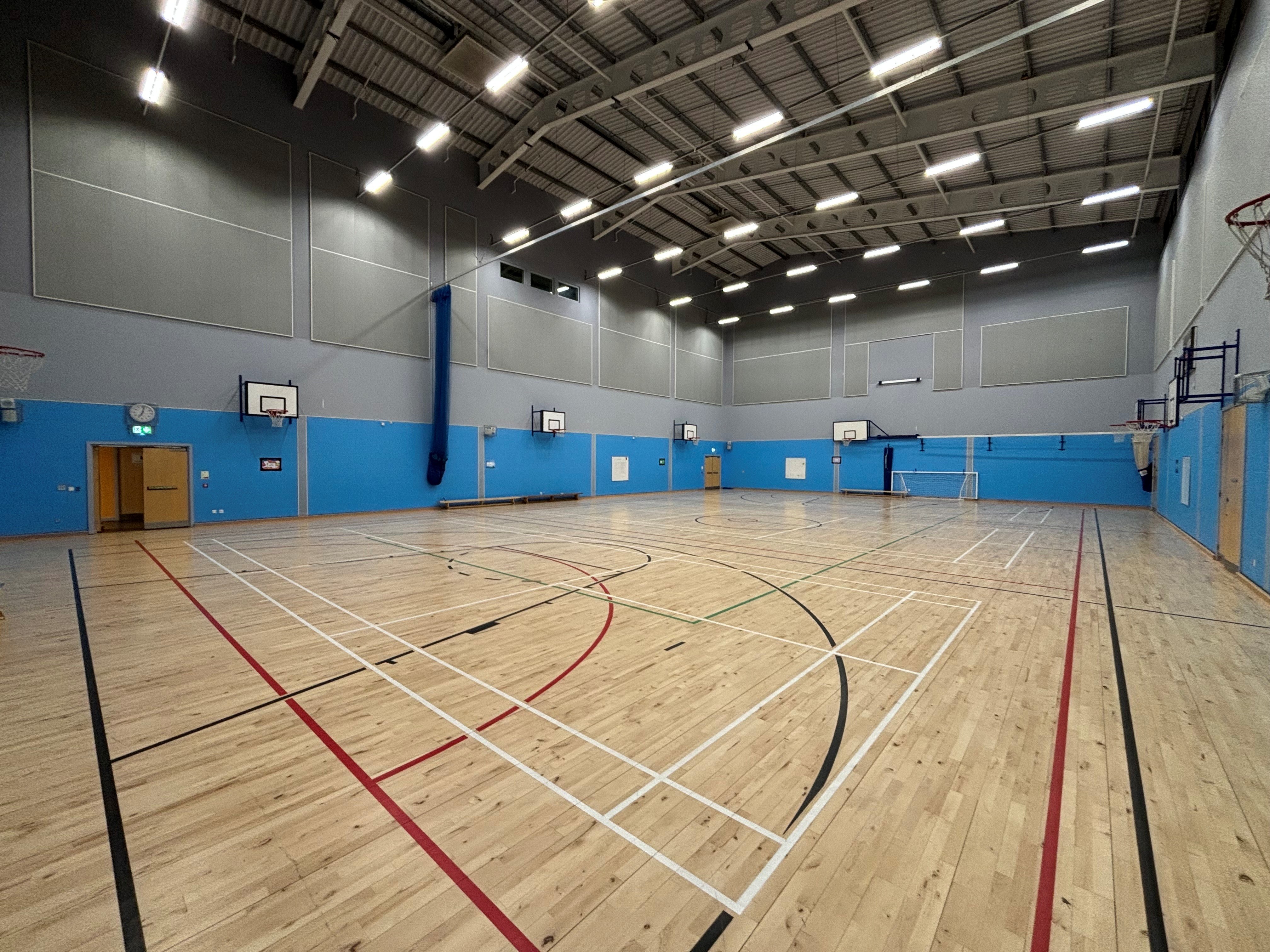 A spacious games hall featuring both badminton court and basketball markings on the floor and basketball hoops affixed to each wall.