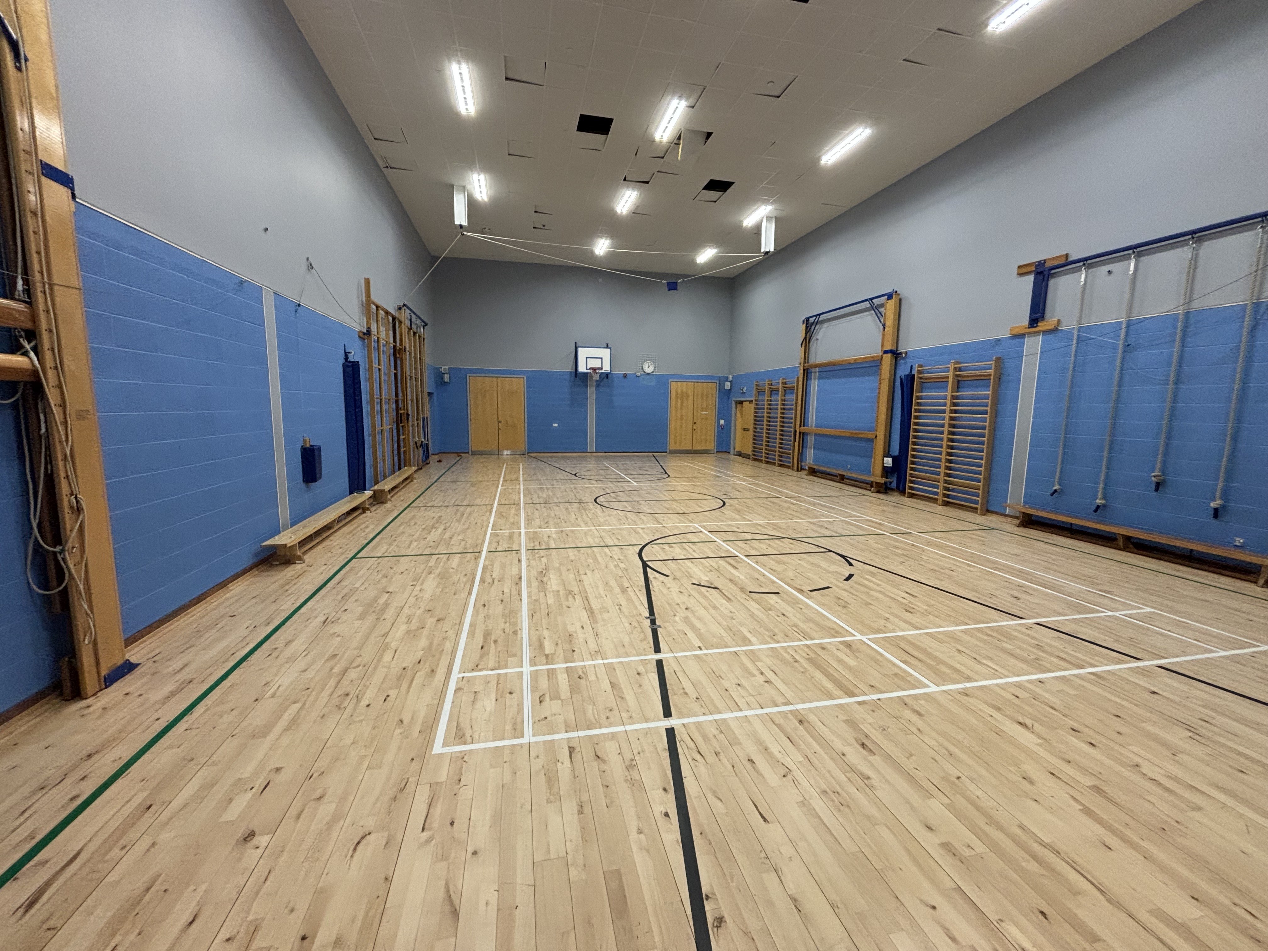 A spacious gymnasium, equipped with a badminton court and marked for basketball, complete with hoops mounted at each end. Additionally, various gym apparatus are positioned along the side walls.