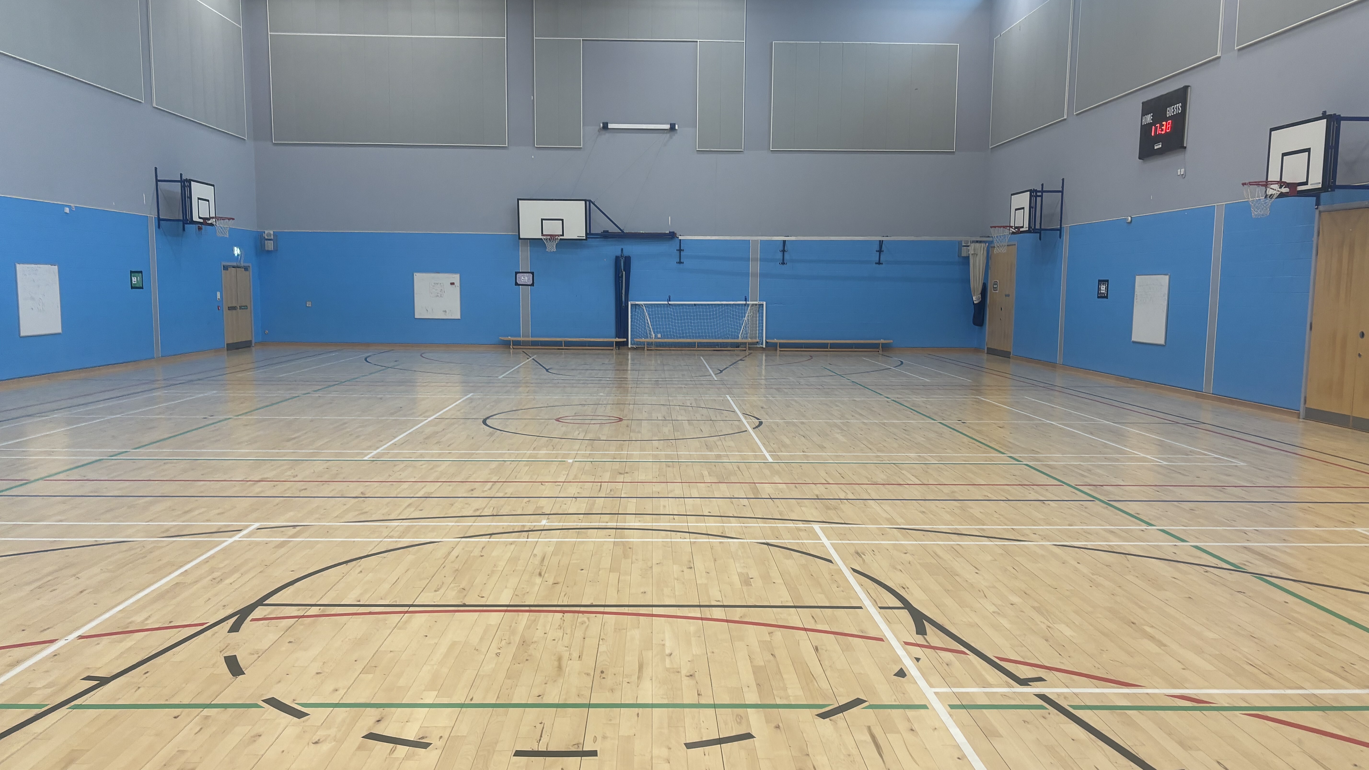 Large sports hall with wooden flooring, wall-mounted basketball hoops and football goal. Painted floor markings for badminton, basketball and multi-sports.