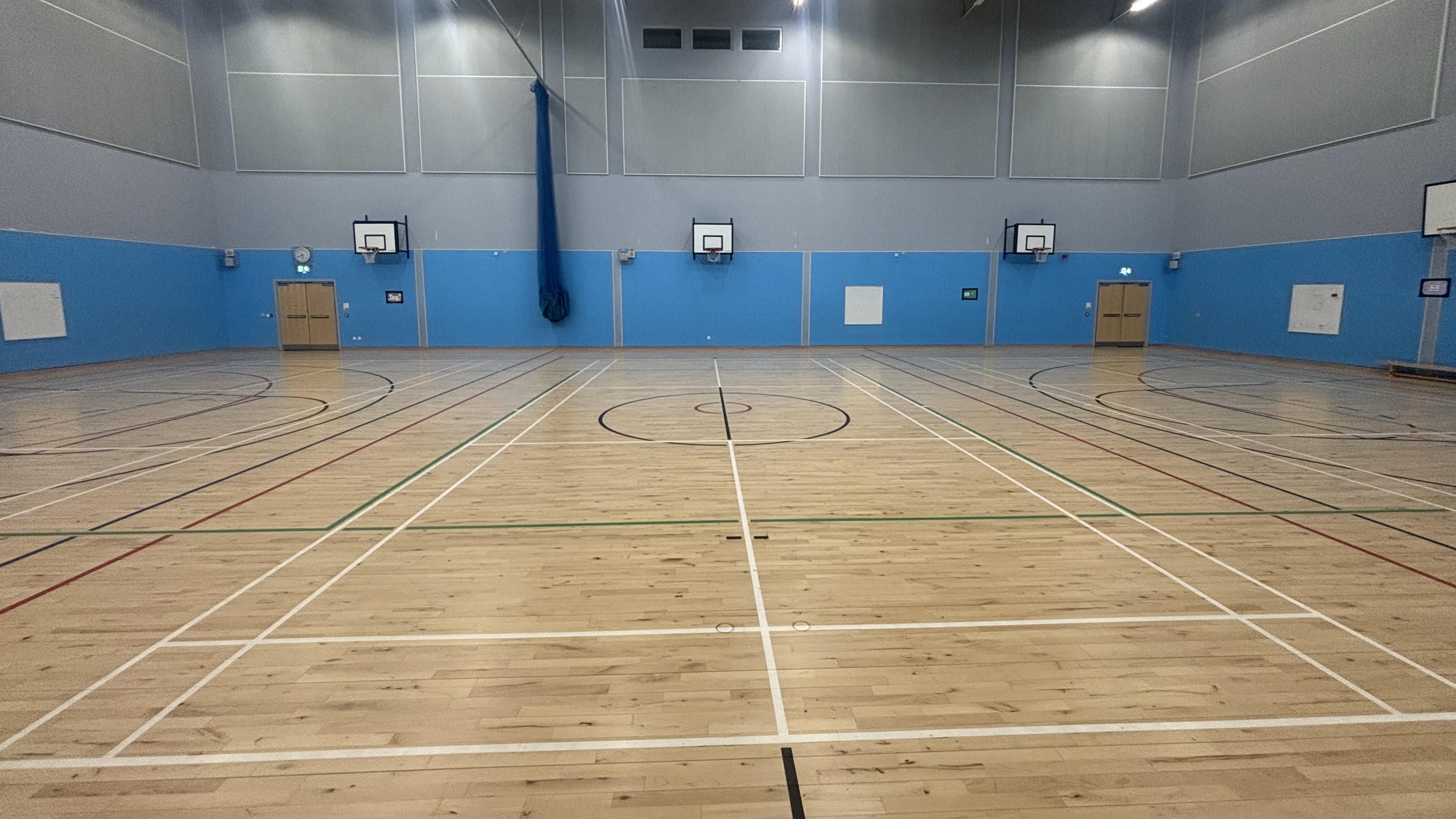 Large sports hall with wooden flooring, wall mounted basketball hoops and painted floor markings for badminton, basketball and multi-sports.