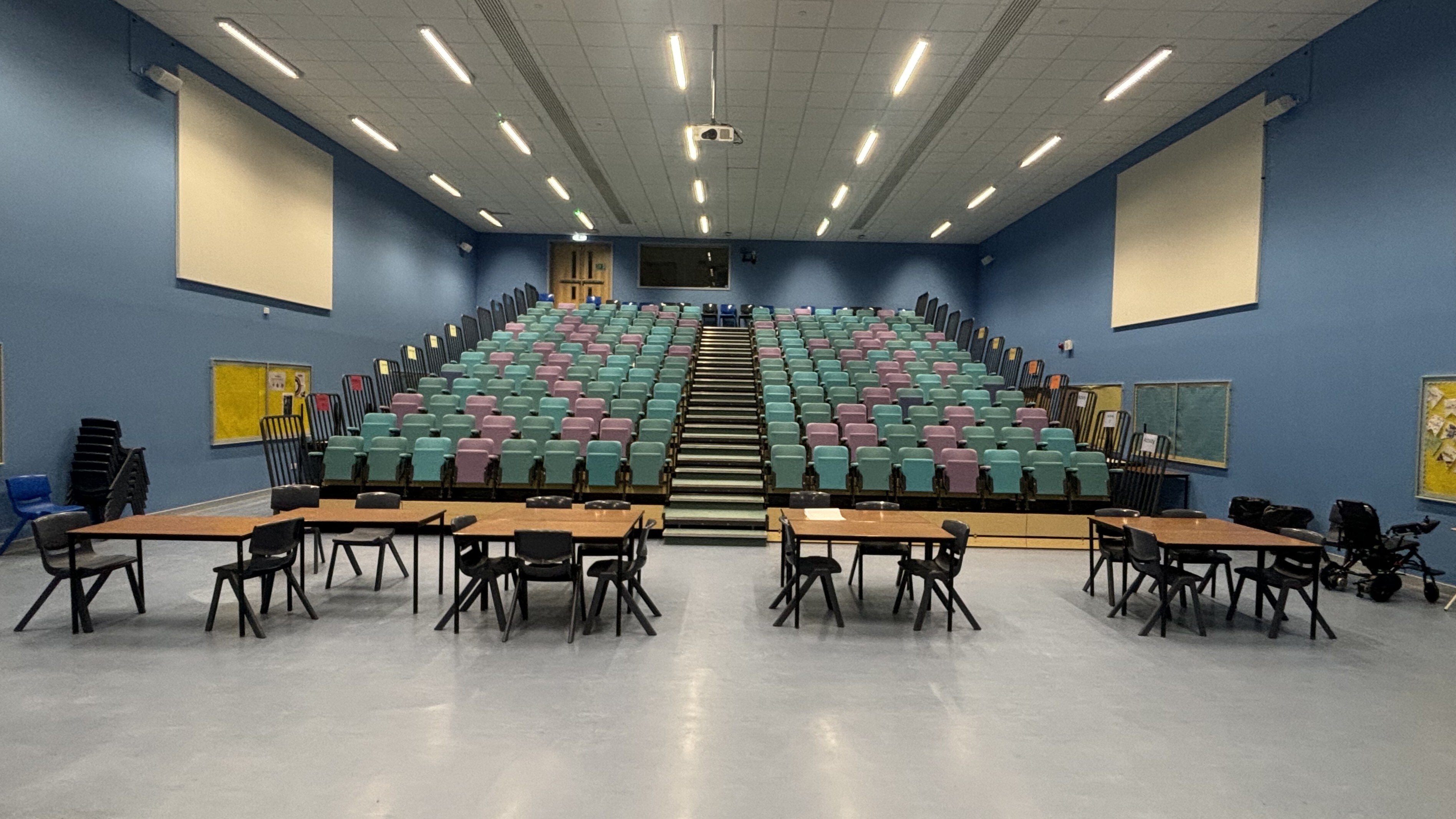 Assembly hall with coloured seating from the bottom of the bleachers looking up. Tables and chairs set up at the bottom across floor area.