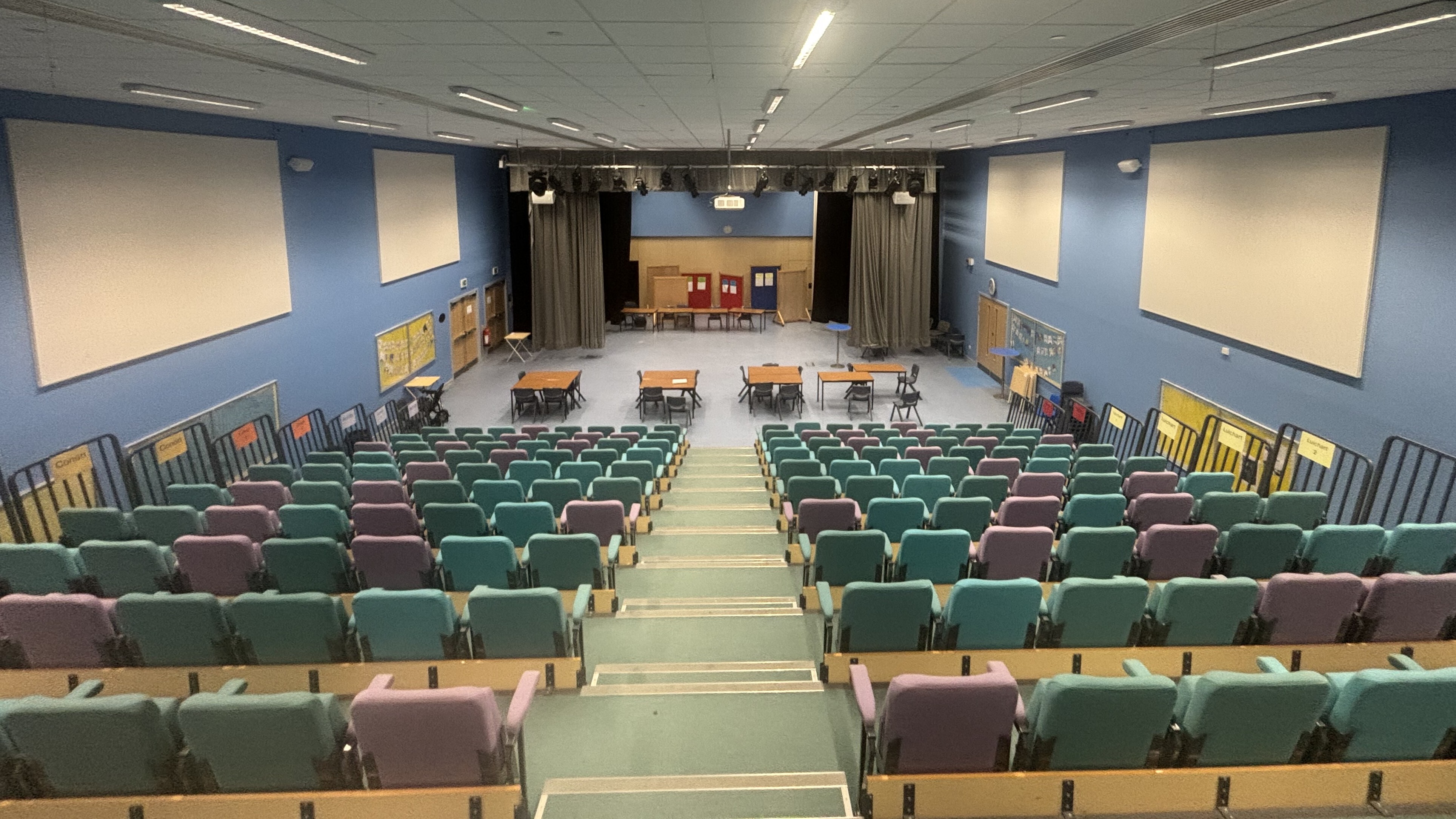 Assembly hall with coloured seating from the top of the bleachers looking down. Tables and chairs set up at the bottom across floor area.