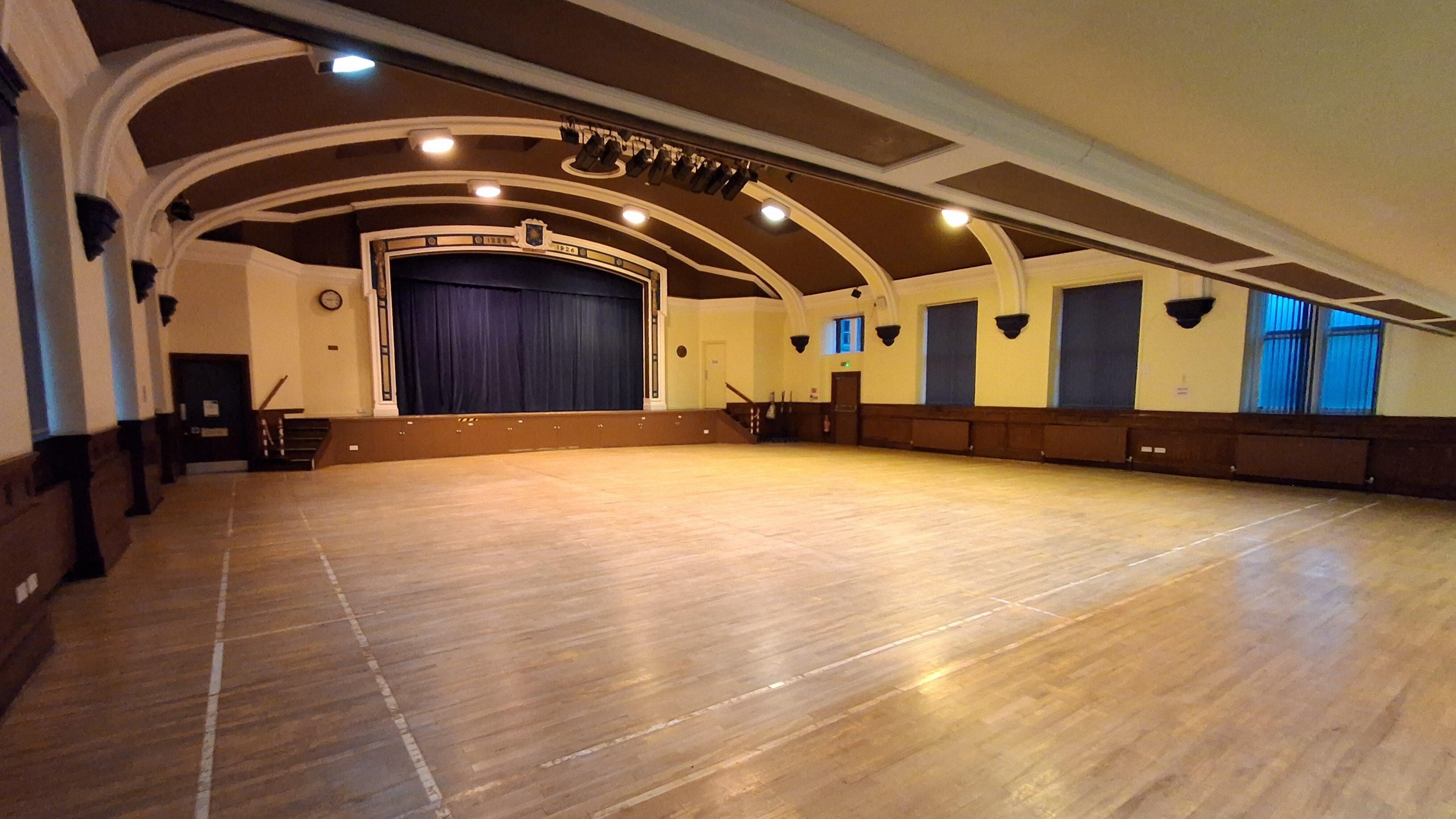 Large hall with wooden flooring. Centre stage at the front with large blue curtain and stairs either side up to the stage. Doors either side.