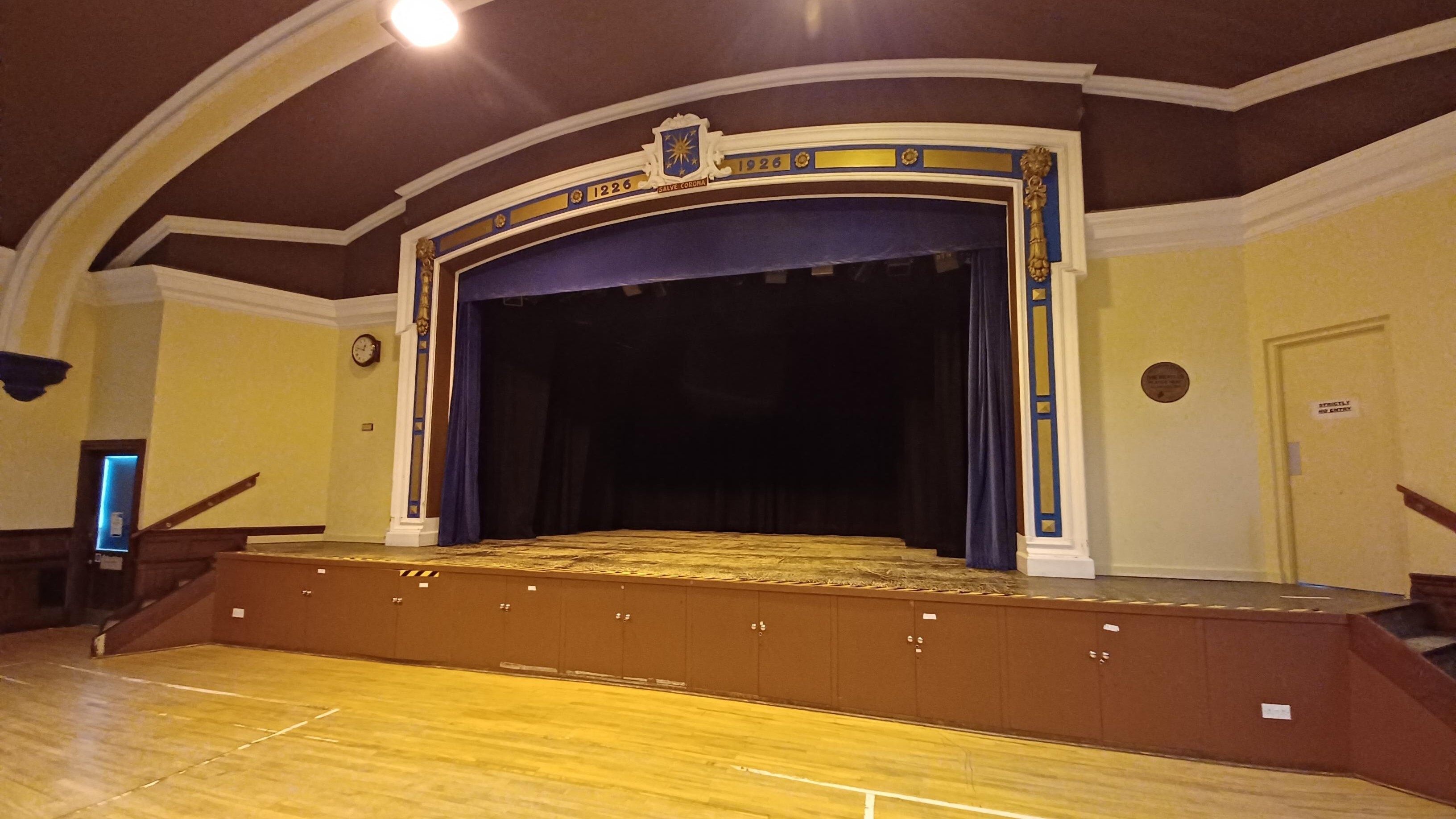 Large hall with wooden flooring. Centre stage at the front with large blue curtain open and stairs either side up to the stage.
