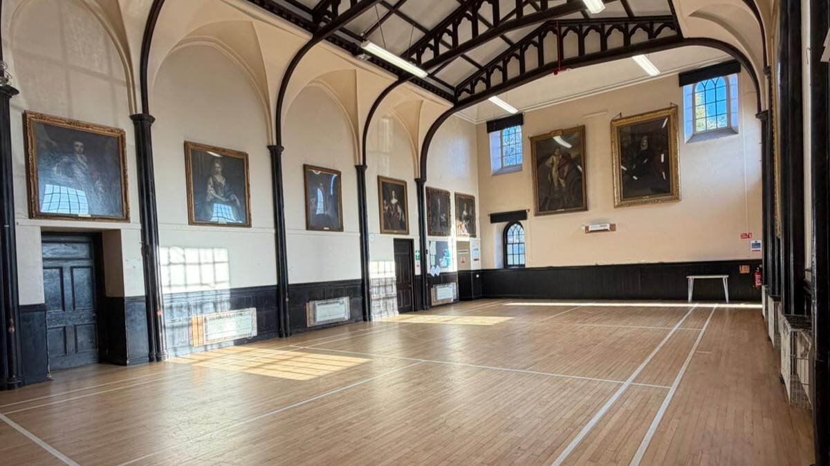 Large hall with arched roof painted black. Several large portrait paintings have been displayed on the walls. Wooden flooring with badminton court painted lines.