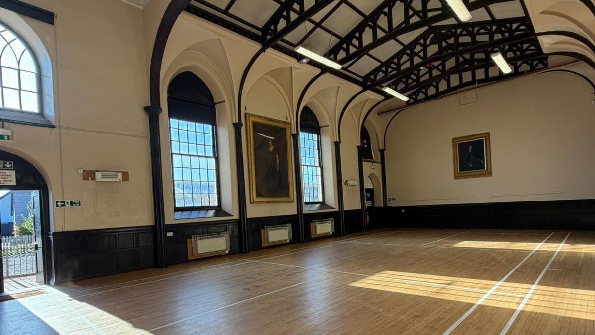 Large hall with arched roof painted black and large glass windows. Several large portrait paintings have been displayed on the walls. Wooden flooring with badminton court painted lines.