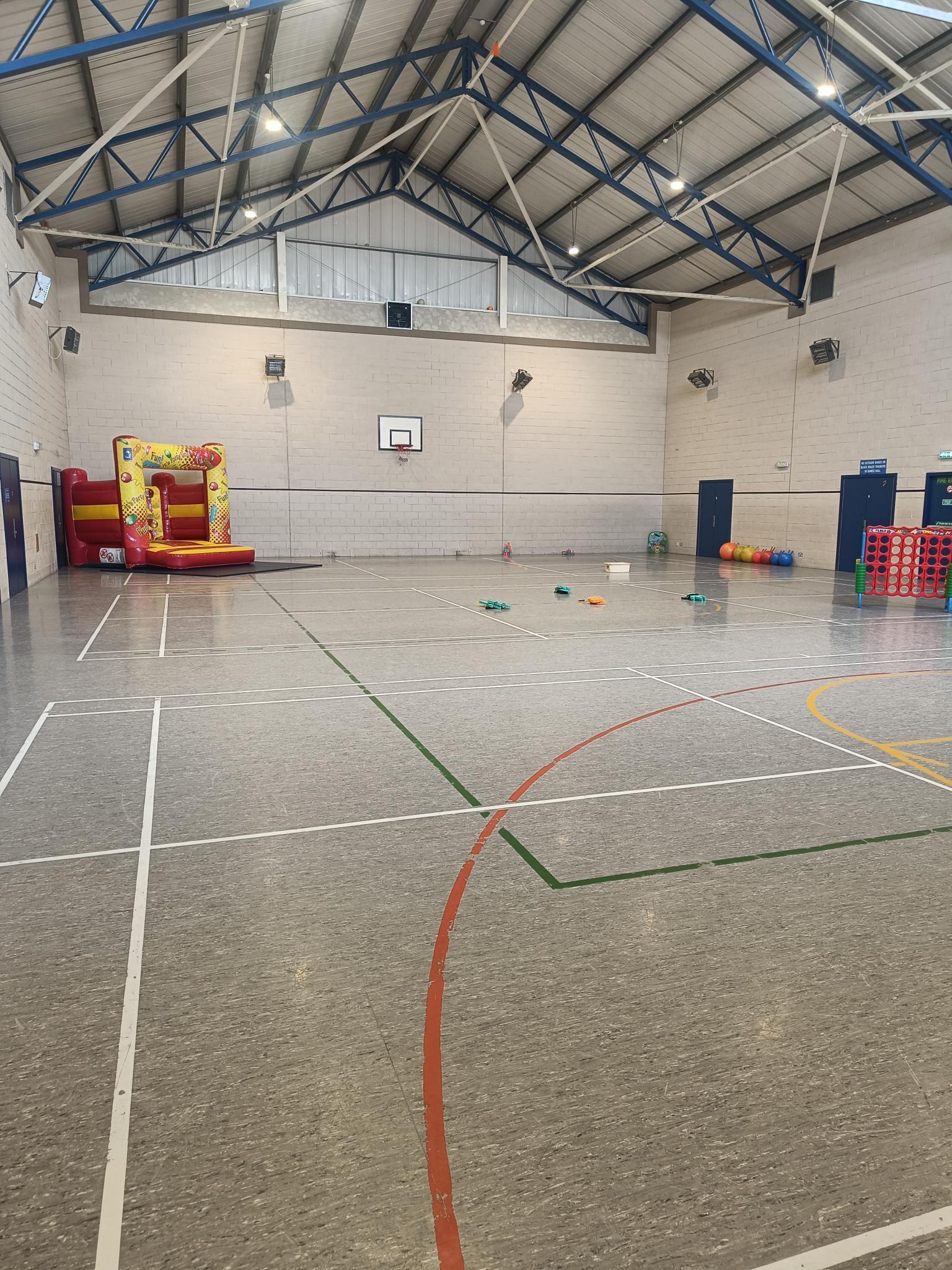 The games hall features floor markings designated for badminton and basketball, along with a basketball hoop positioned on the end wall and a bouncy castle in the corner.