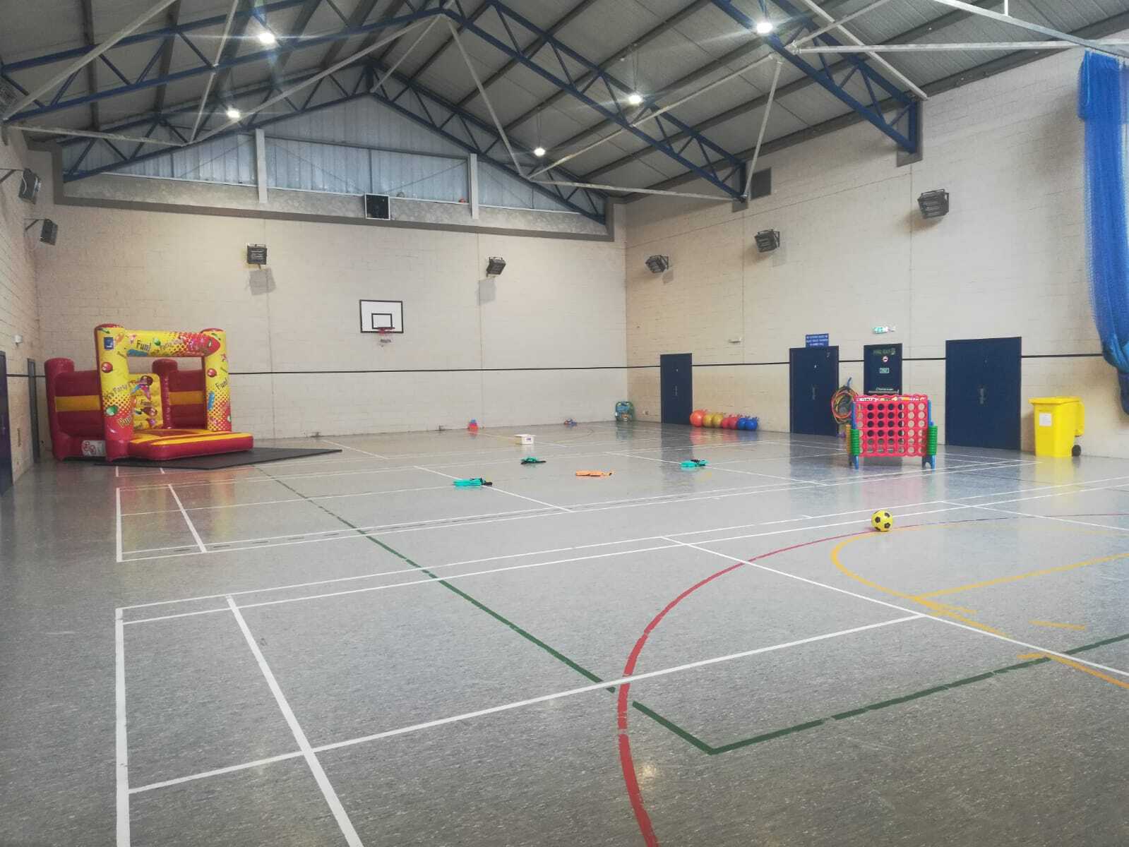 The games hall features floor markings designated for badminton and basketball, along with a basketball hoop positioned on the end wall and a bouncy castle in the corner.