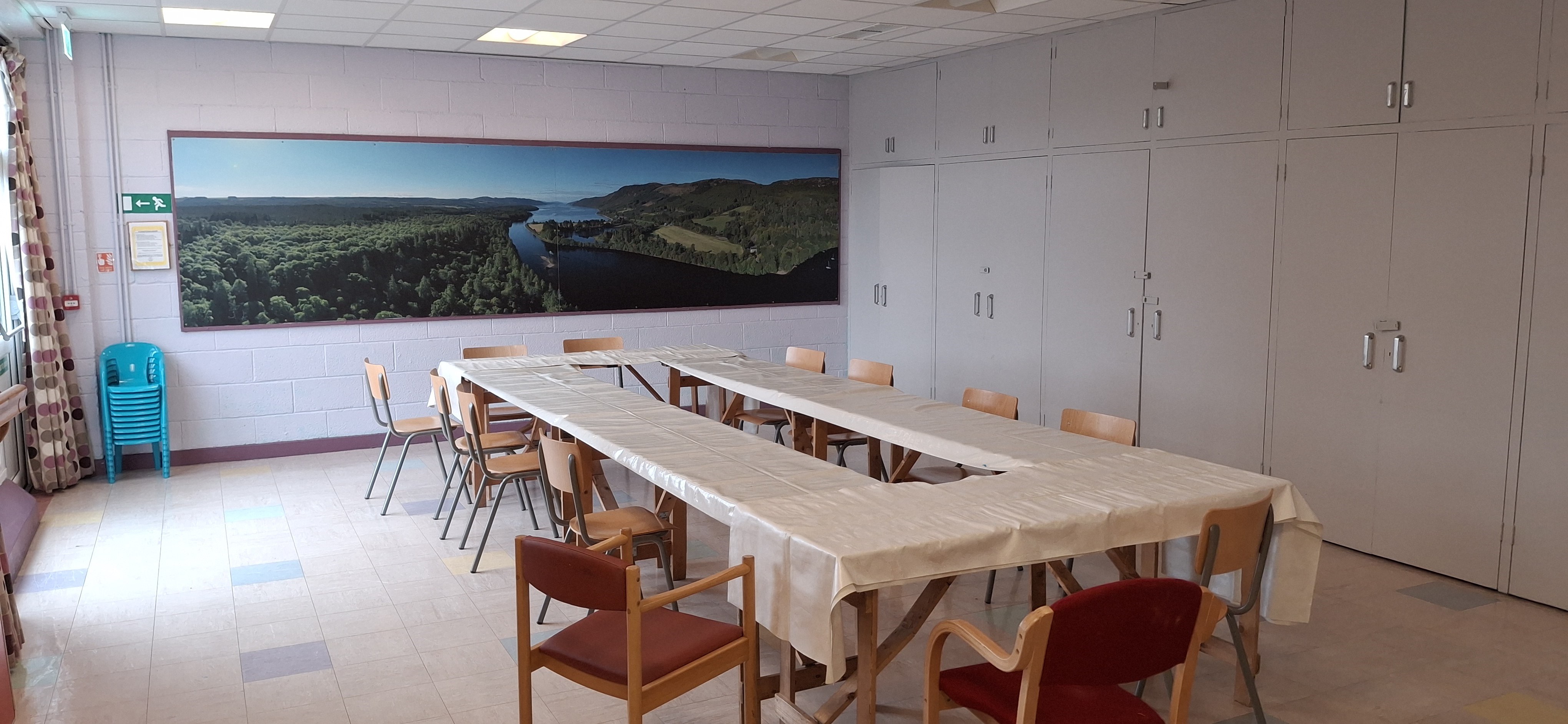 A spacious room showcases a beautiful scenic picture on the far wall, with tables covered in protective plastic and surrounded by chairs, making it ideal for children's activities.