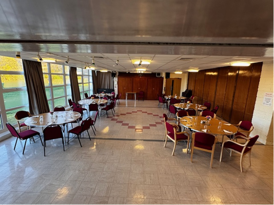 A spacious room featuring floor-to-ceiling windows and patterned linoleum flooring, arranged with circular tables complemented by chairs, making it an ideal setting for lunch / dinner events.