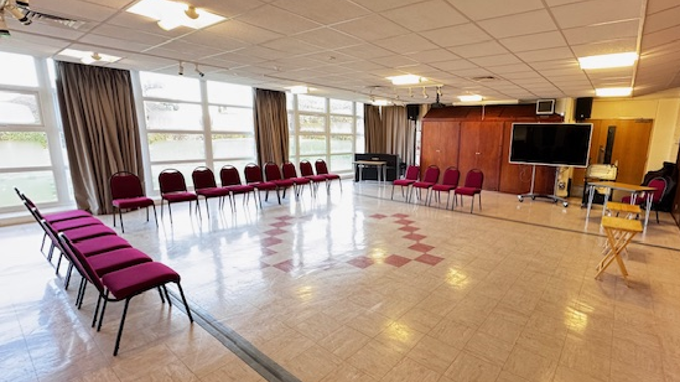 Large room with polished floor and glass windows to the left. Chairs set up in a semi-circle with portable tv and tables at the front.