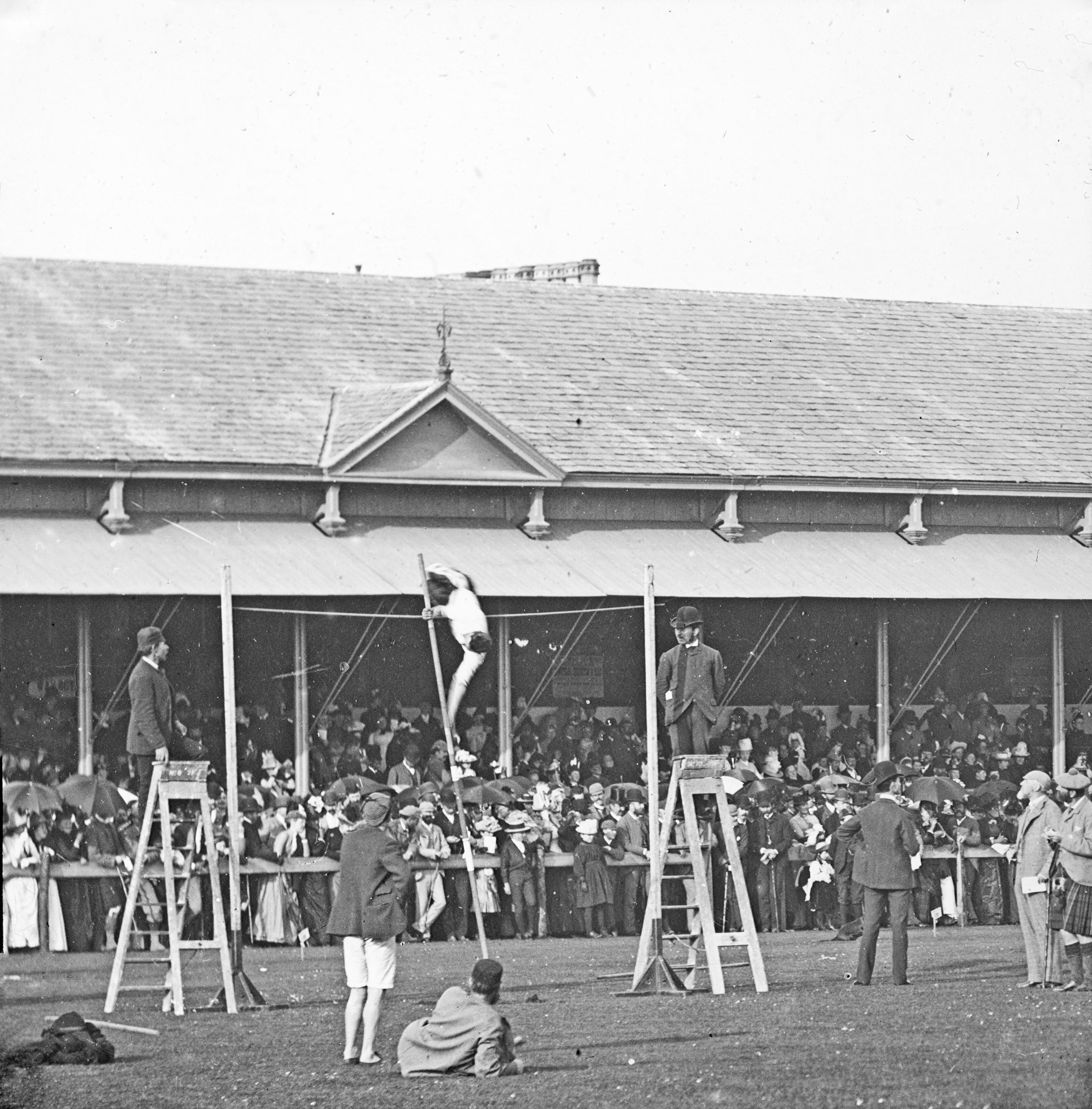 Black and white photograph of a competitor pole vaulting at the Northern Meeting Games in the early 1900s, with spectators watching in the background.