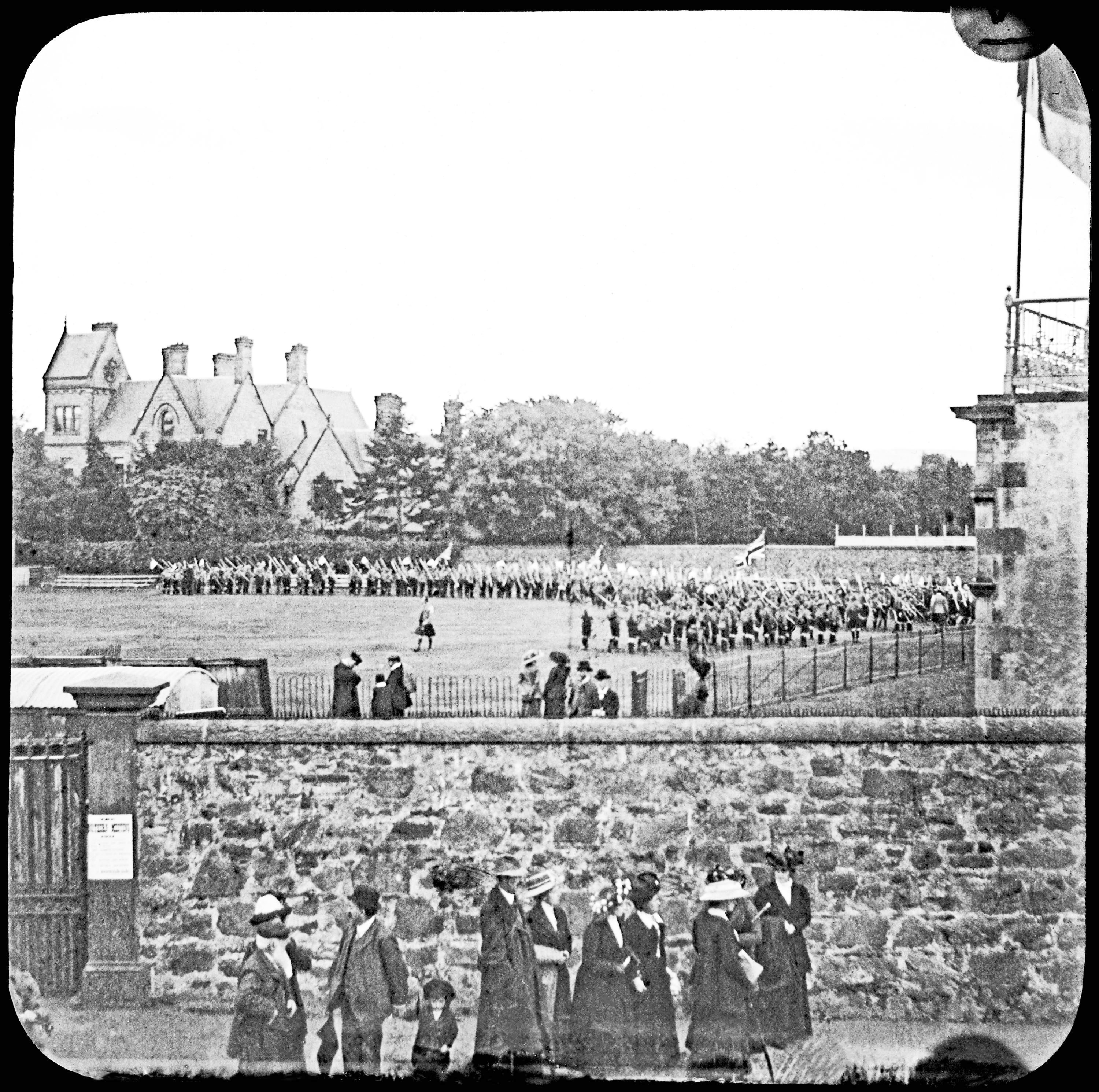 Black and white lantern slide photograph viewed from outside the boundary wall of Northern Meeting Park, showing a large procession of Scouts assembled on the field beyond, with spectators lining the perimeter and a Victorian villa visible in the background, c.1911.