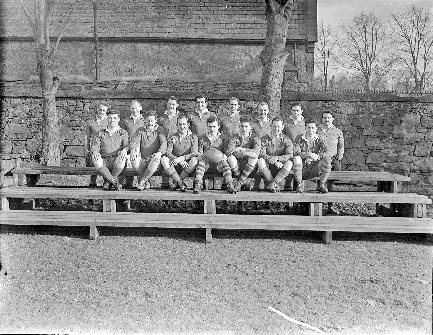 Black and white photograph of a rugby team of fifteen players posed in two rows on wooden benches against a stone wall, wearing matching jerseys, with one player holding a ball, date unknown.