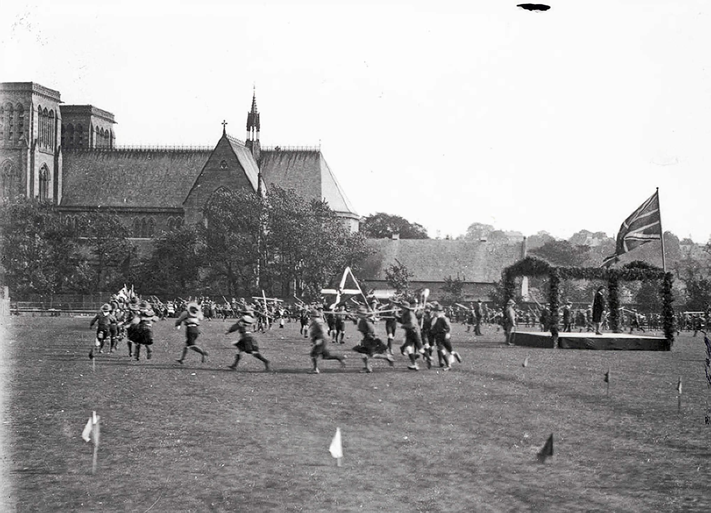 Black and white photograph of uniformed Scouts running across Northern Meeting Park during a skills display, with Inverness Cathedral and a decorated Union Jack-draped platform visible in the background, c.1911–13.