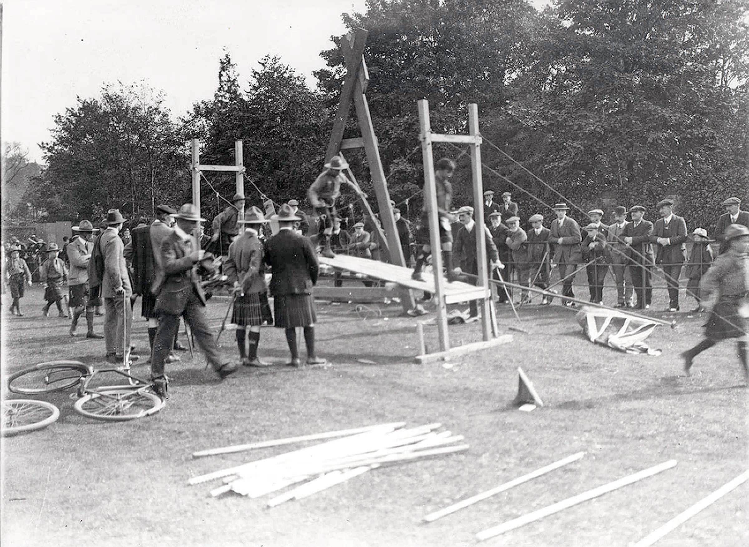 Black and white photograph of Boy Scouts erecting a wooden frame structure on the field at Northern Meeting Park, watched by a crowd of spectators, as part of a public display of Scoutcraft skills, c.1911–13.