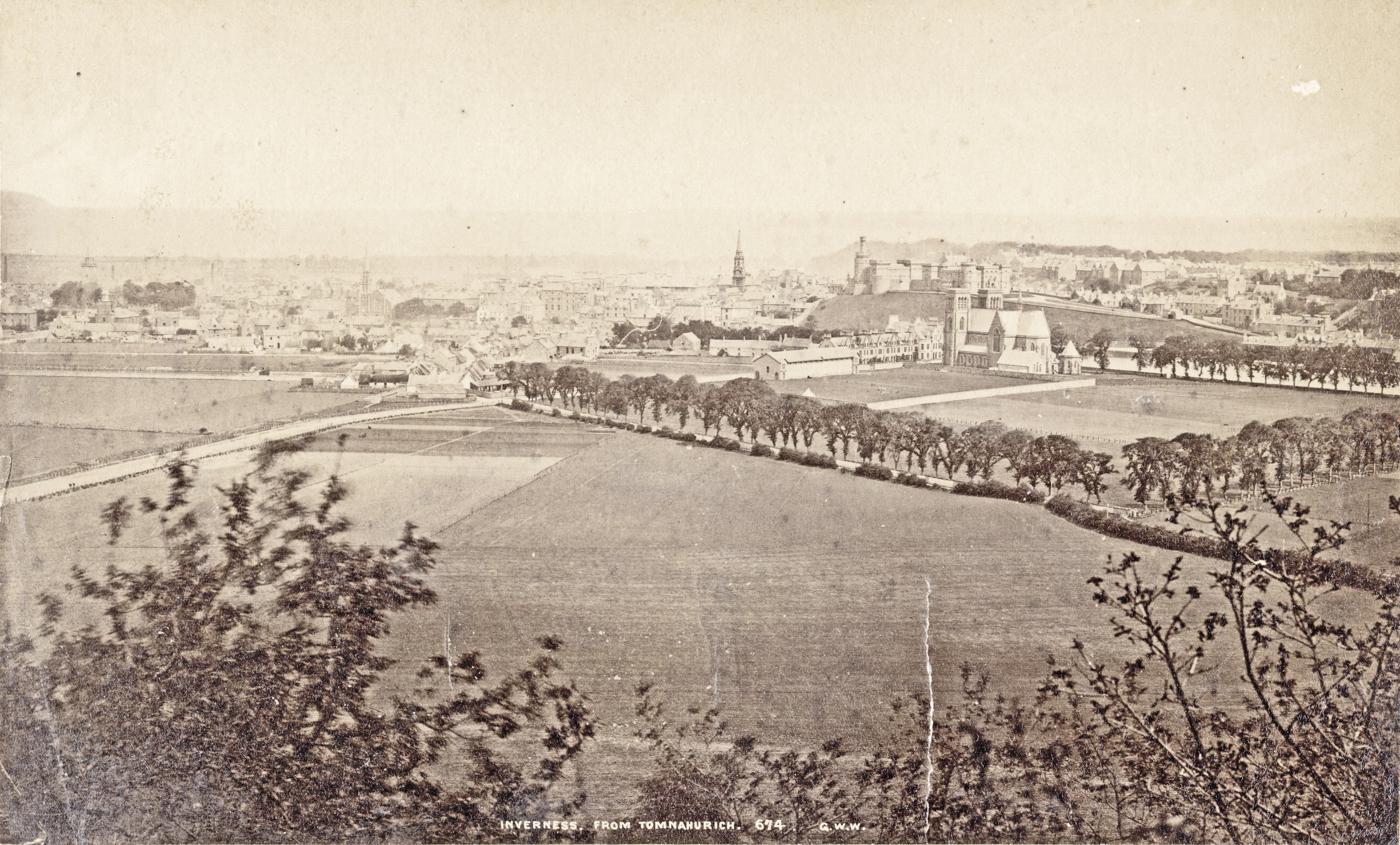 Sepia photograph taken from Tomnahurich looking across open fields towards the Northern Meeting Park beside Inverness Cathedral, with Inverness Castle visible on the skyline. The surrounding area is largely undeveloped.