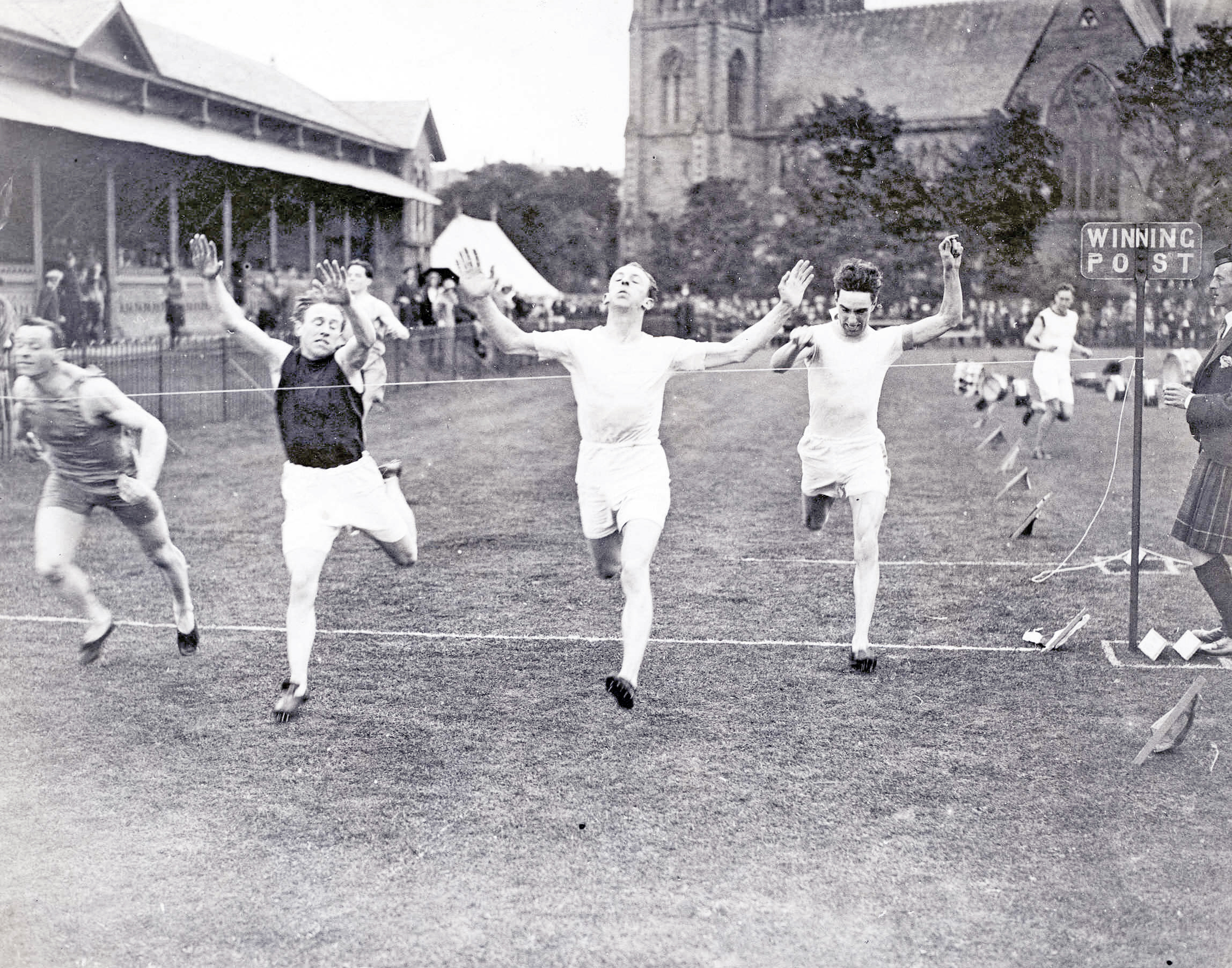 Black and white photograph of athletes crossing the finishing line of the 220-yard sprint at the 1922 Northern Meeting Games, arms raised, with a Winning Post sign to the right.