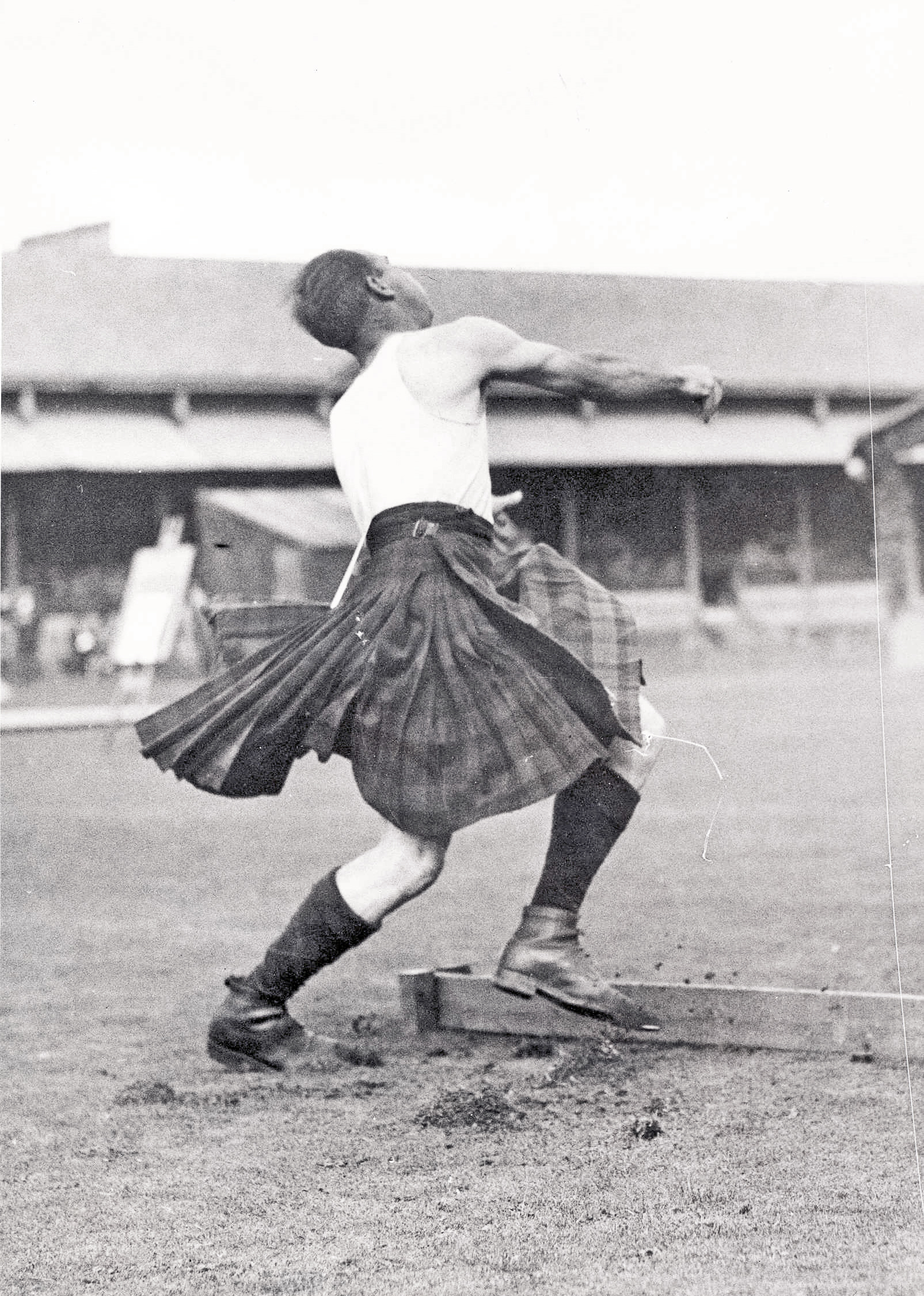 Black and white photograph of a kilted athlete in mid-throw during the hammer throwing competition at the 1936 Northern Meeting Games, with the grandstand in the background.