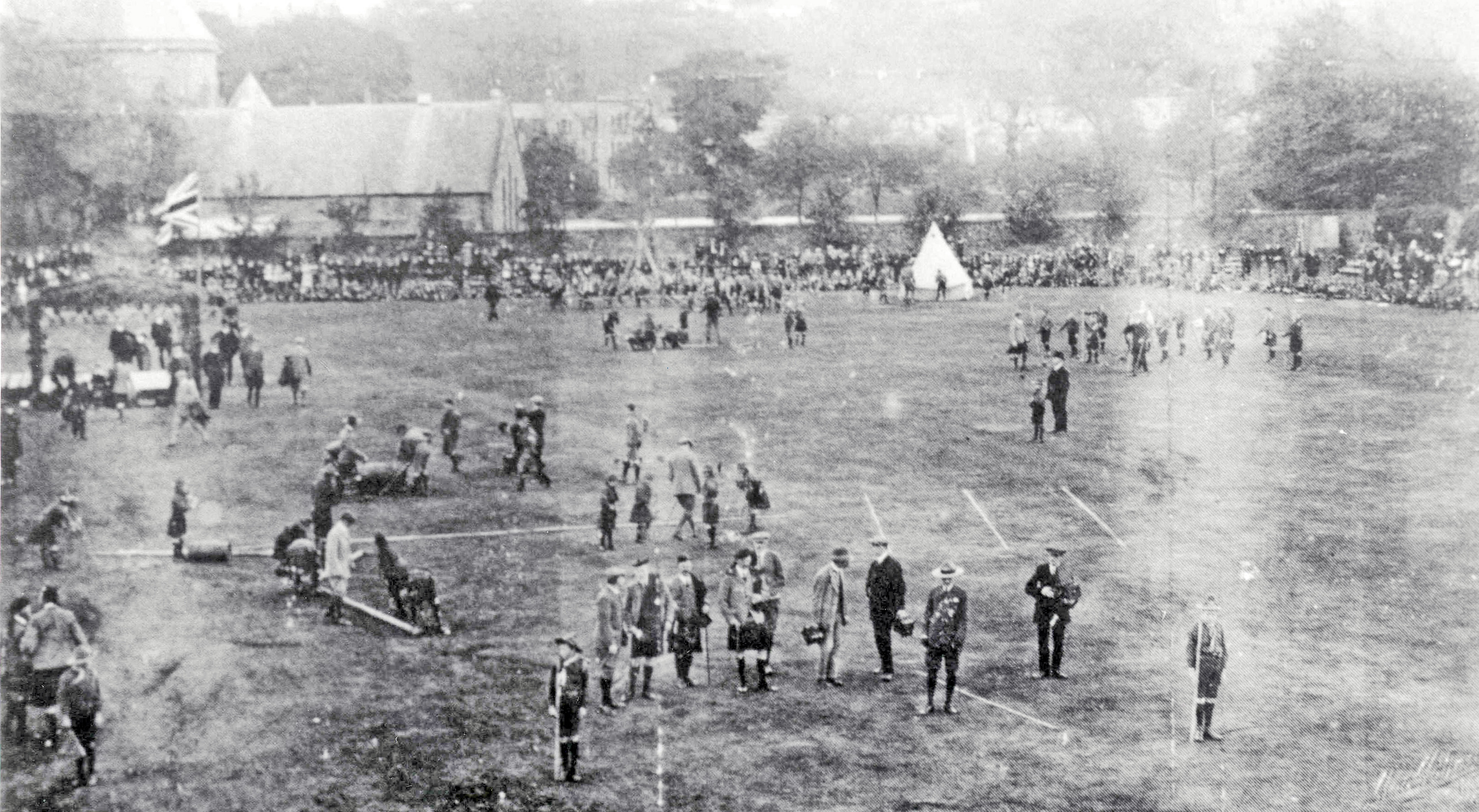 Black and white photograph taken from an elevated position showing hundreds of Scouts and spectators spread across Northern Meeting Park during the 1911 rally, with a large crowd lining the perimeter, a Union Jack flying, and a marquee tent visible in the background.