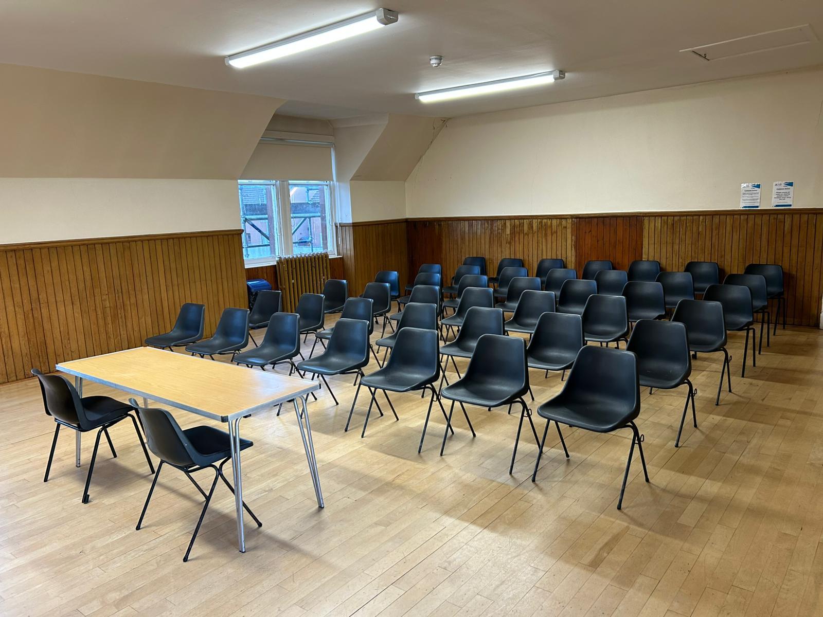 A hall featuring hardwood flooring and abundant natural light that pours in through the windows. The room is laid out with a table and chairs in theatre style.