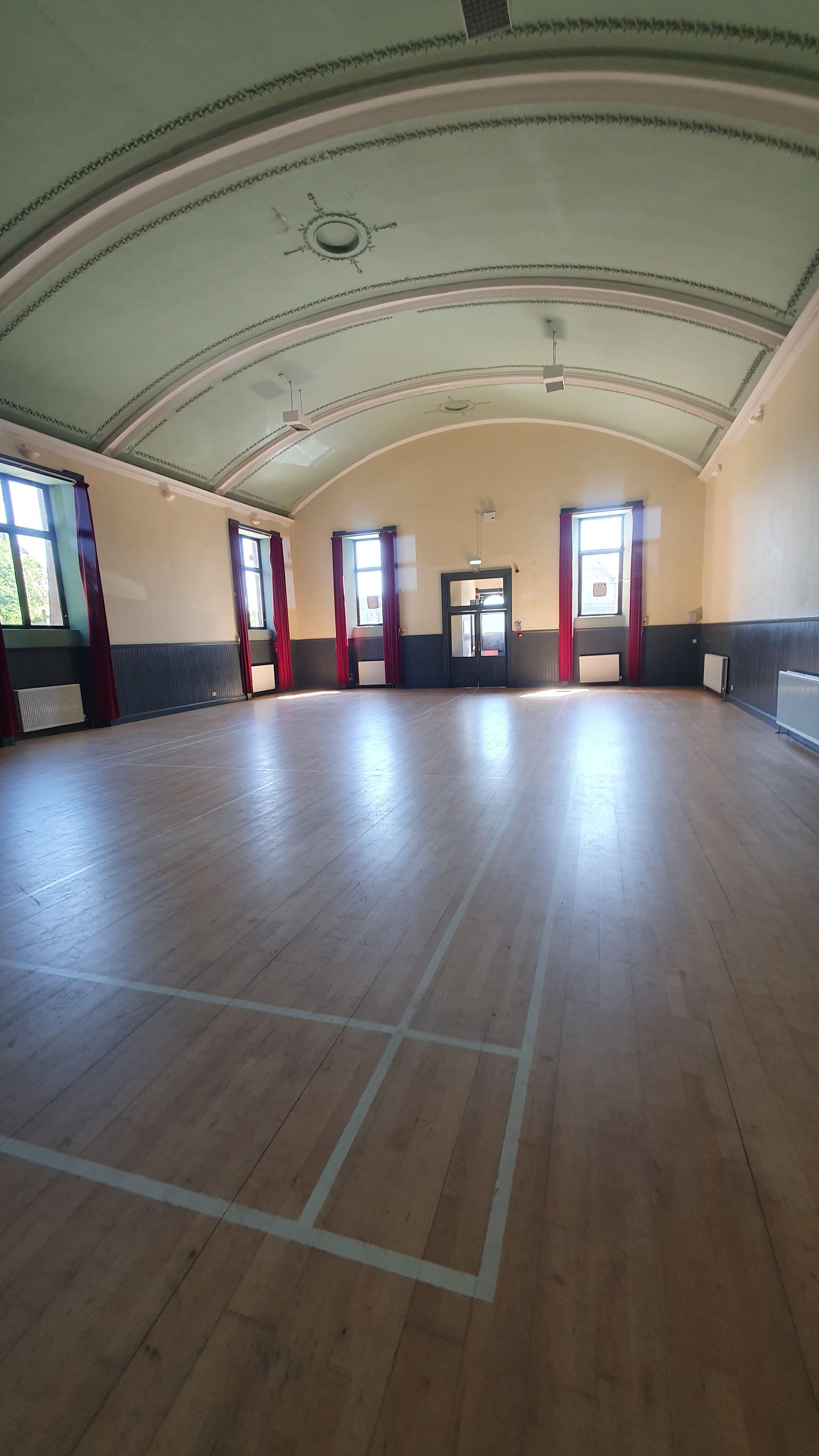 The Hall features a badminton court and is illuminated by natural light that pours in through expansive, curtained windows. Its elegantly arched ceiling contributes to the overall ambiance, creating a warm and inviting atmosphere.