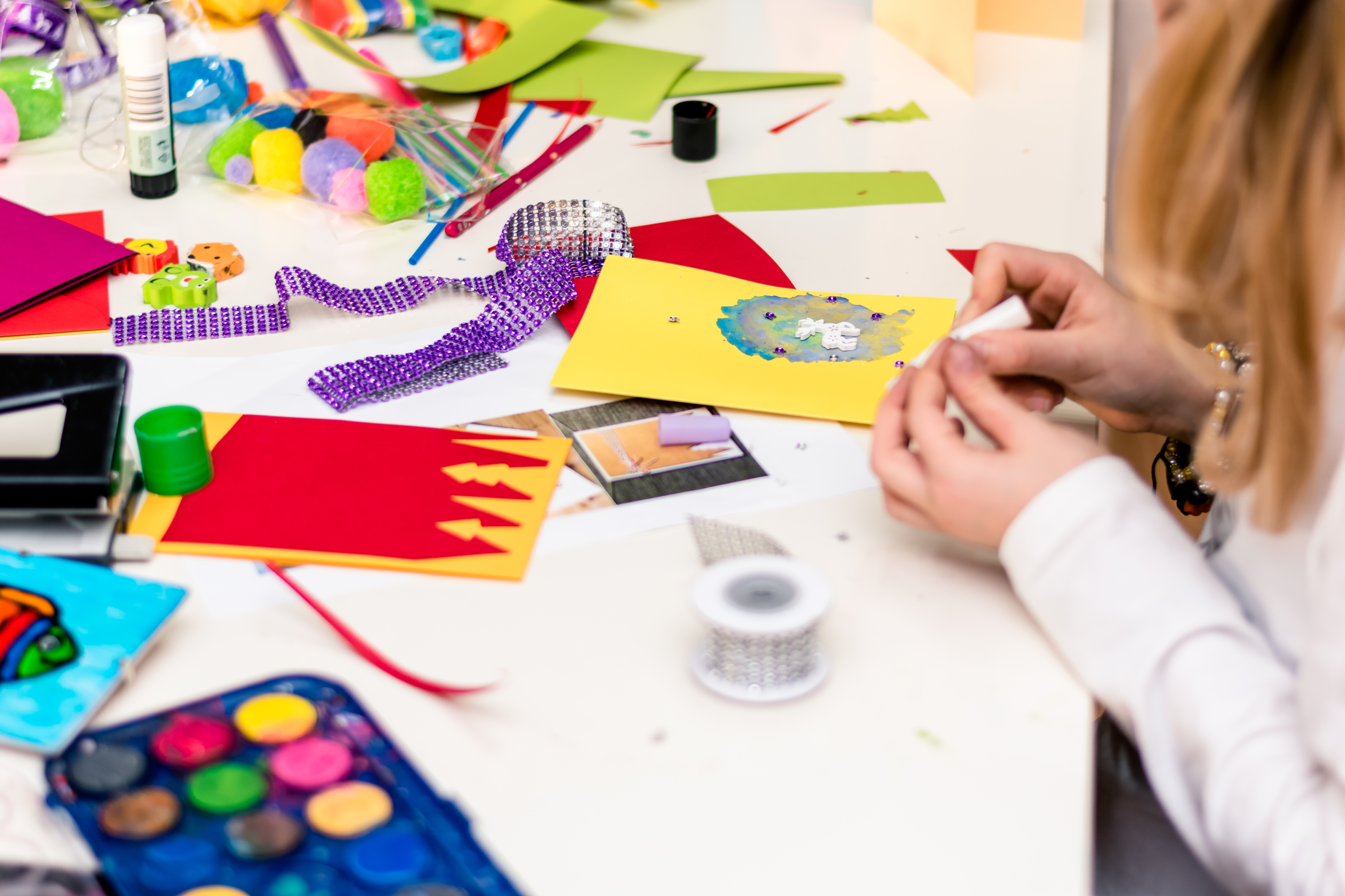Teenage girl sitting at table doing arts and crafts. Coloured paper and accessories scattered across table, as well as glue sticks and paint sets.
