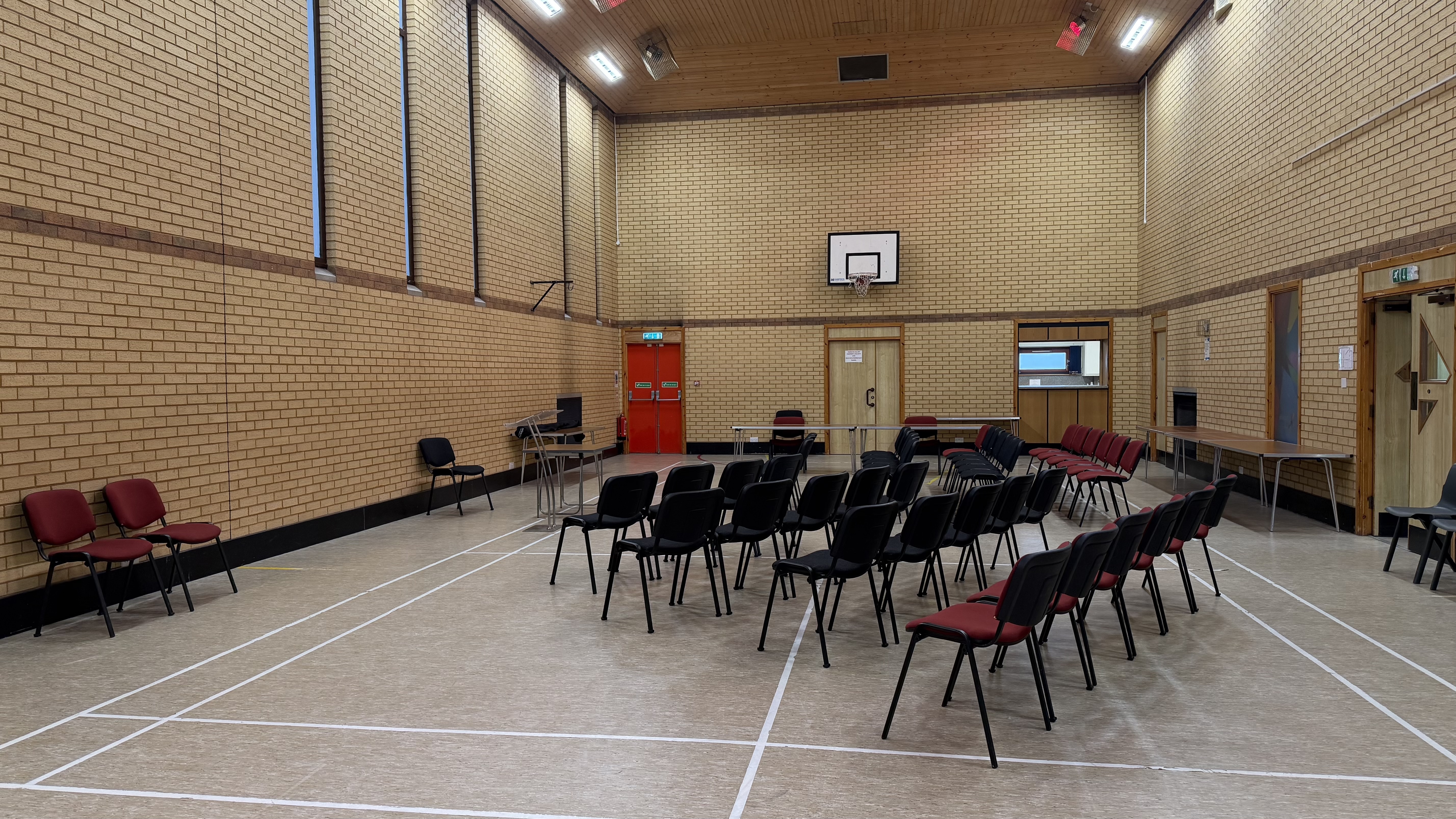 Rows of black and red soft chairs set up in sports hall. Theatre format for public meeting with tables around the outside of hall.
