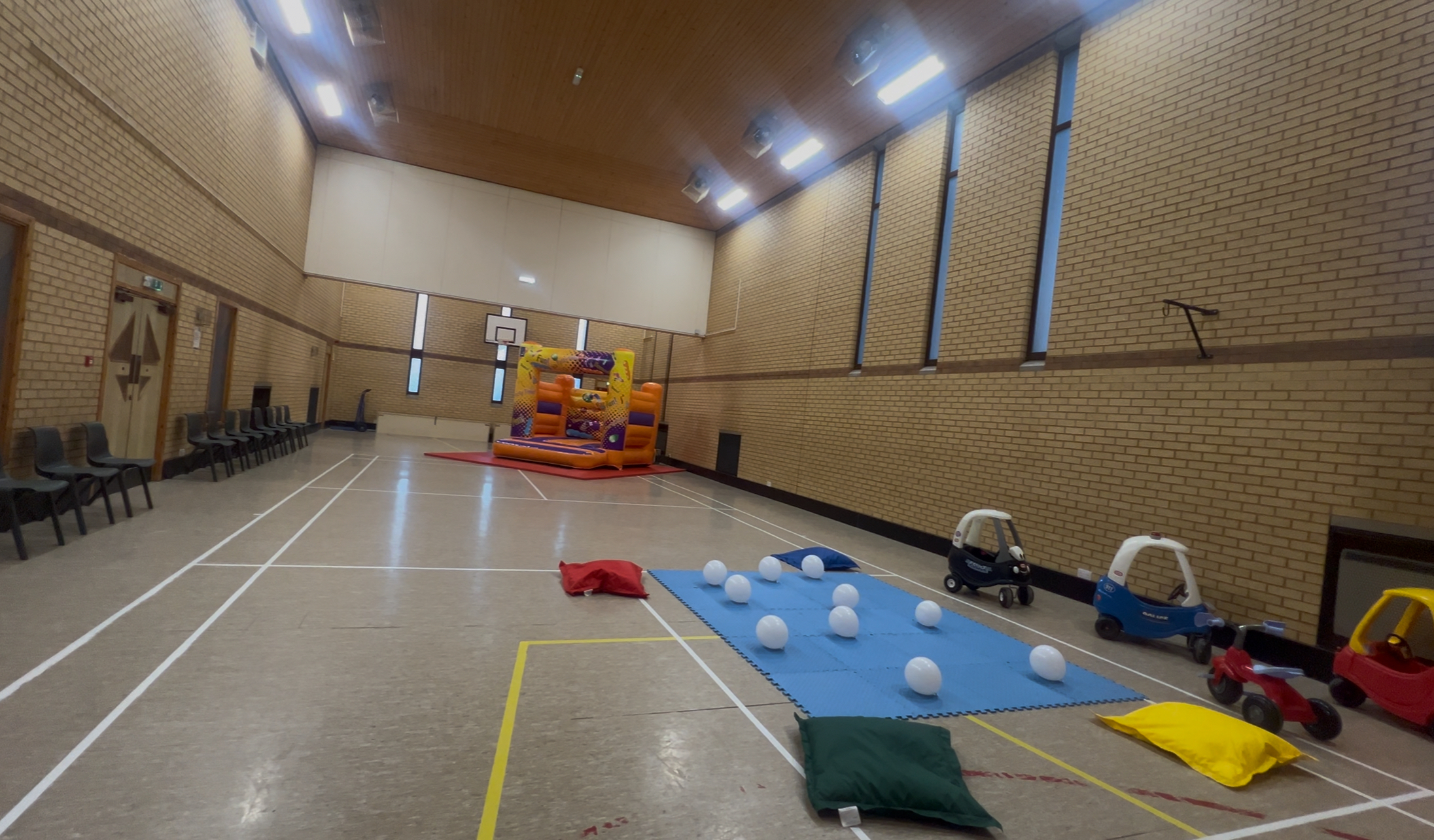 Large hall with children's jigsaw mats laid out in a square with balloons, ride-on cars and beanbags surrounding the area in the foreground. In the background, a bouncy castle is set up towards the back of the hall.