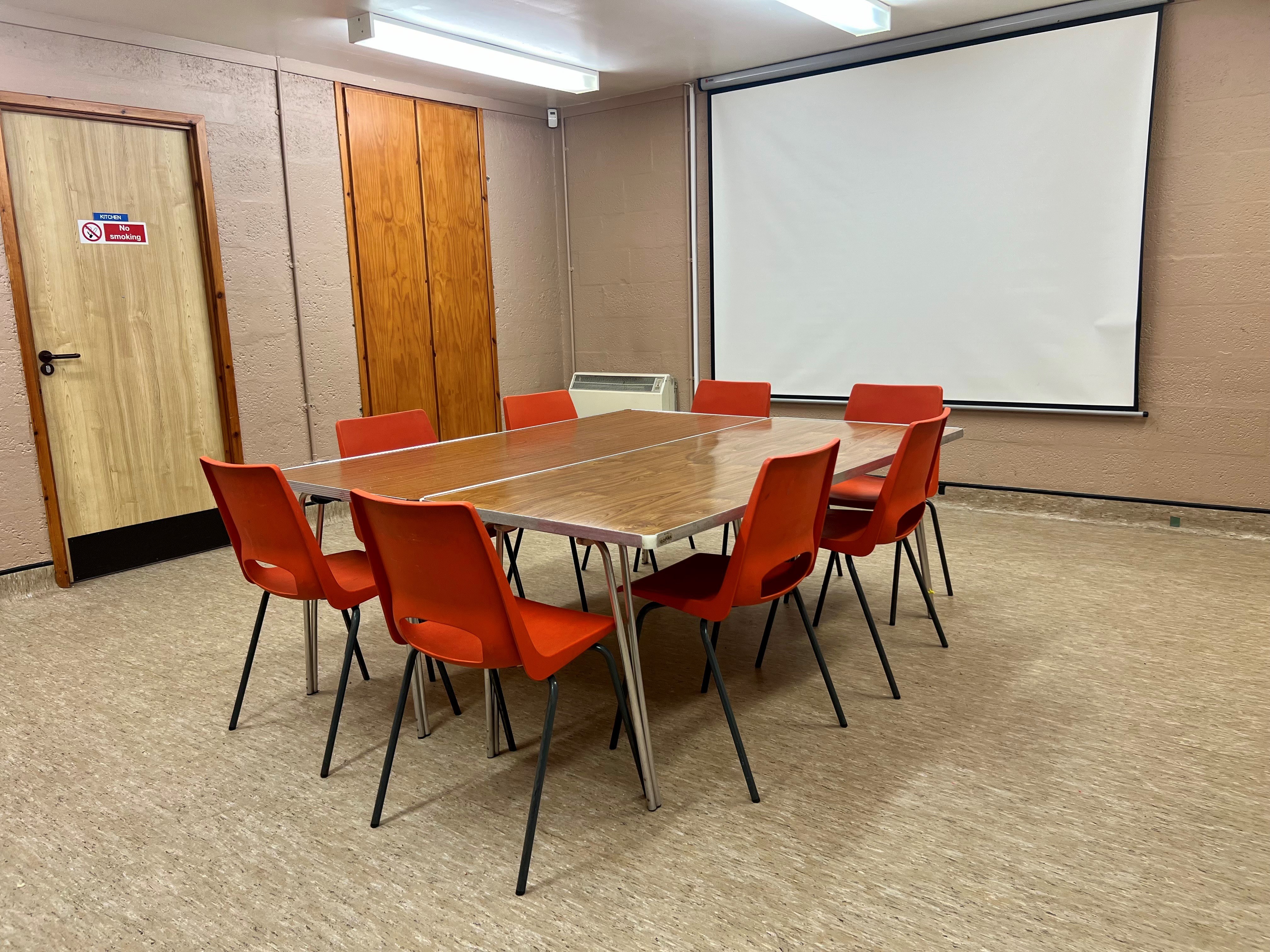 The King Duncans room is a versatile square space that can be configured for various purposes. This photo illustrates a meeting room setup, featuring large tables and surrounding chairs positioned centrally. Additionally, it includes a large wall-mounted white screen, ideal for presentations.