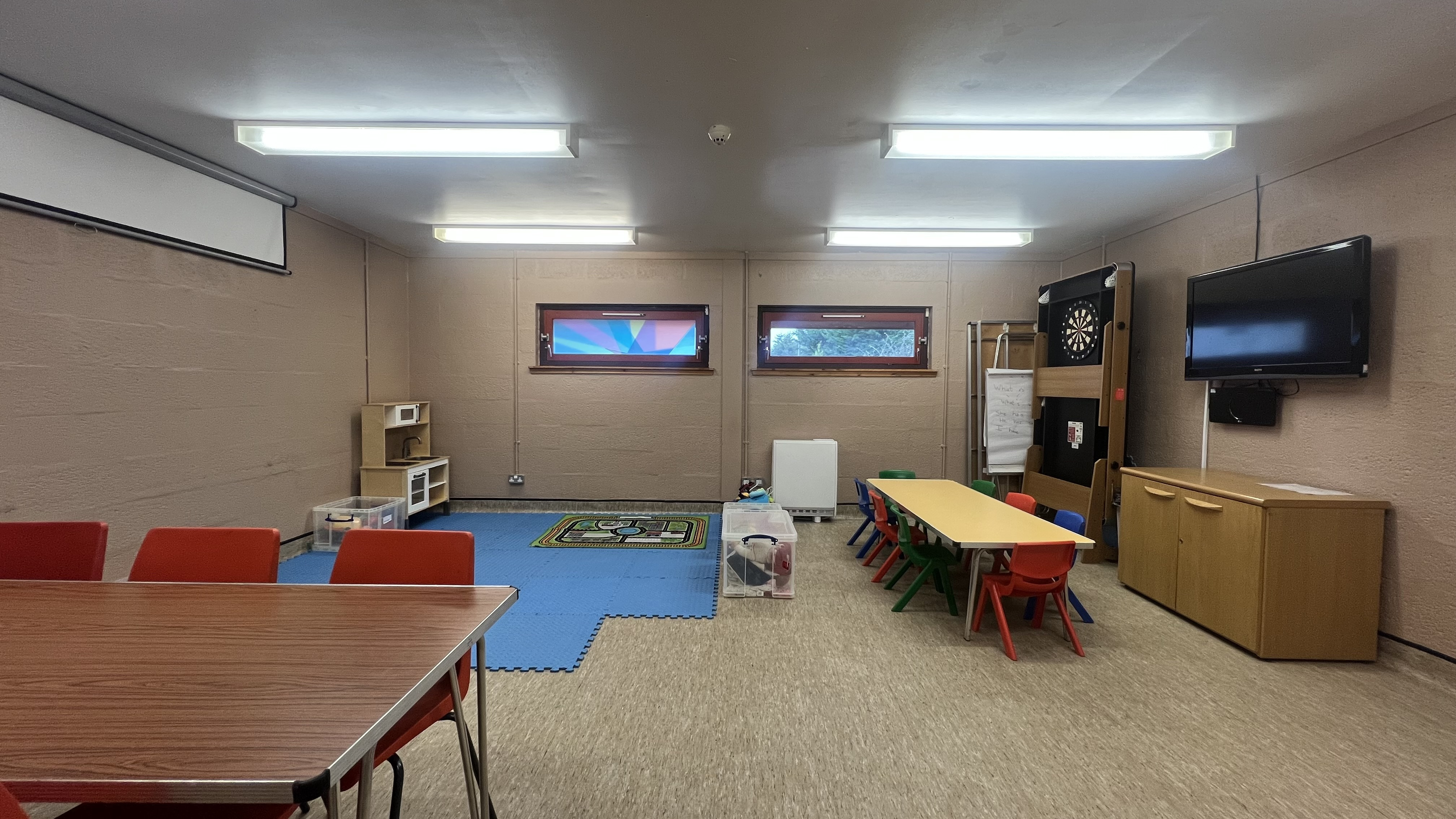 The King Duncan's room is a versatile square space that can be configured for various purposes. This photo shows the room being utilised as a creche, with soft matting covering the floor in the corner, along with child-sized tables and chairs, and a portable dartboard. Additionally, there is a wall-mounted television on one of the walls.