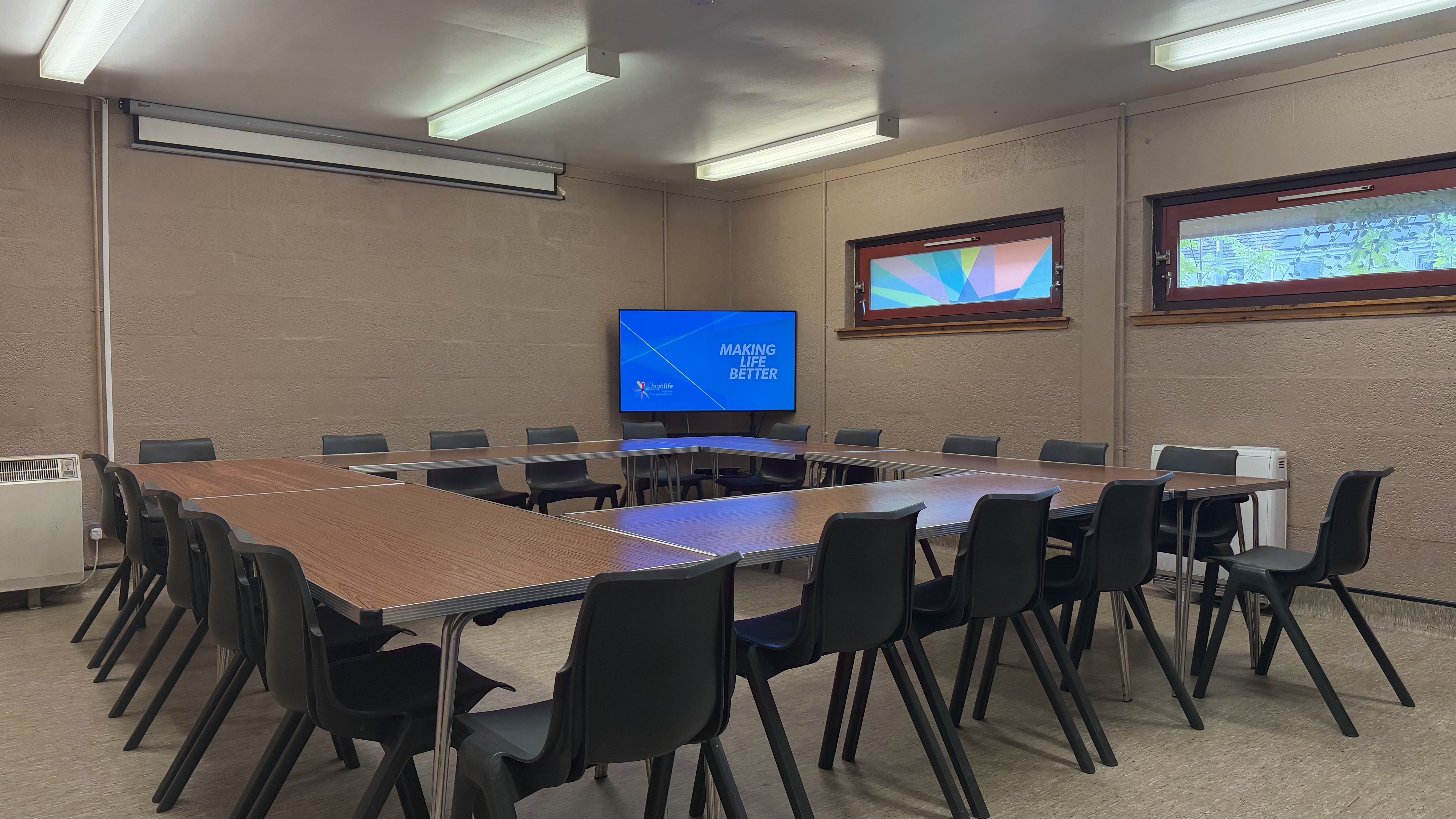 Large meeting set up with tables configured in a large hollow square with 20 chairs around the outside. Large TV mounted on the wall displaying High Life Highland branding with the 'Making Life Better' tagline.