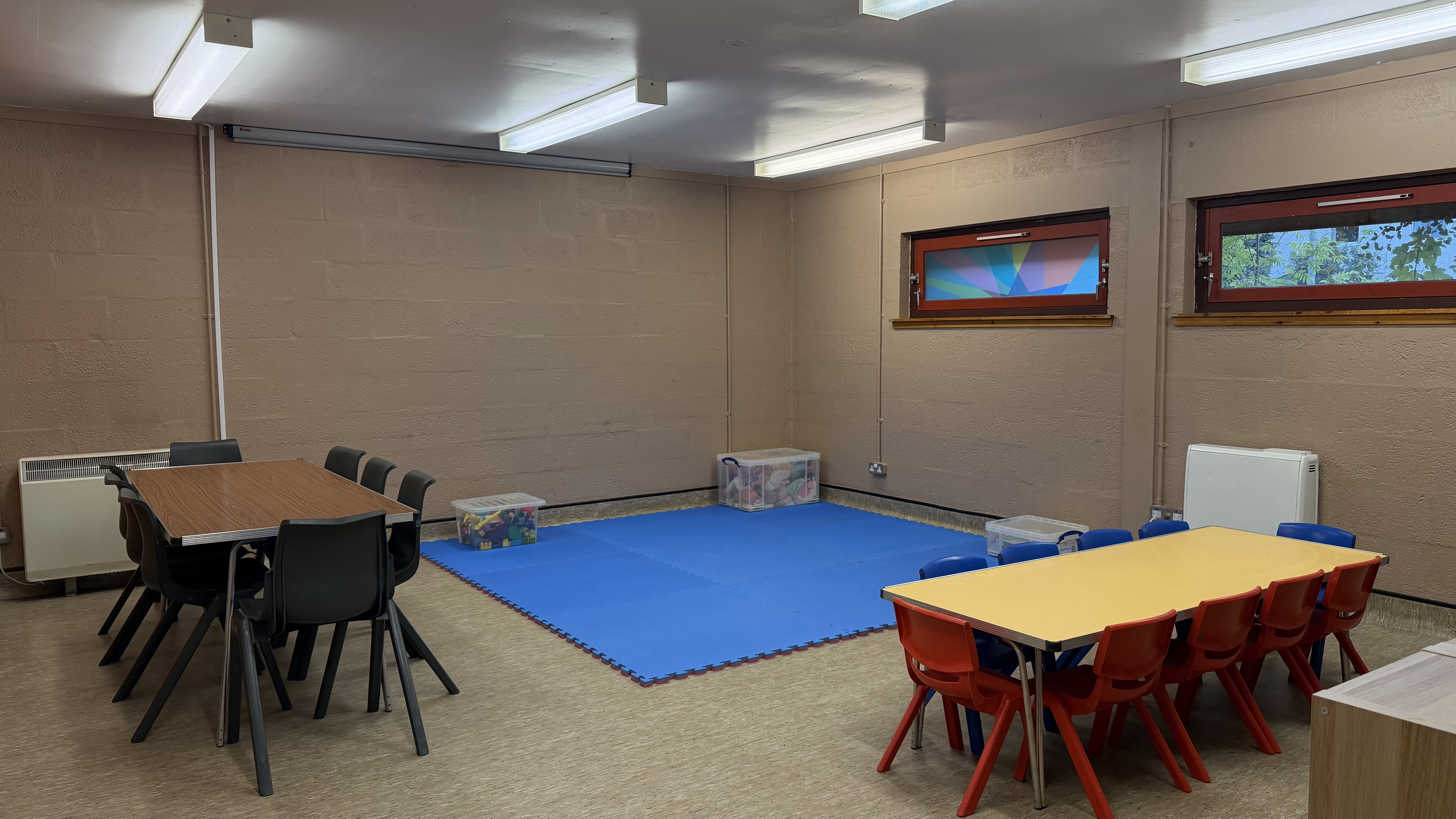 King Duncan's Room set up for a creche, with a blue foam play mat in the centre, storage boxes of toys, a low table with red and green children's chairs, and a separate table with adult chairs along the wall.