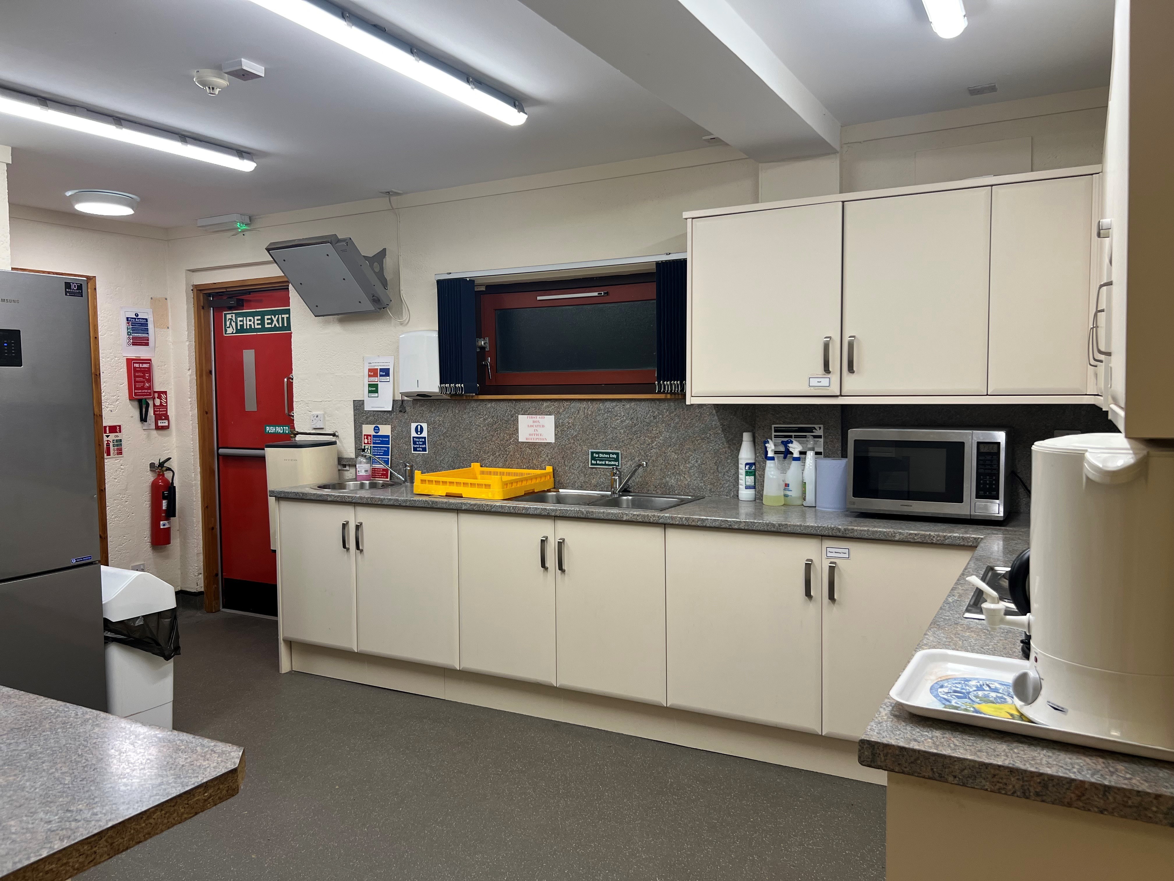 The kitchen is generously sized, featuring an abundance of countertop space and numerous cupboards. It is equipped with a sink, a large fridge-freezer, a microwave, and a kettle. Additionally, there is a serving hatch that connects to the Ashton Room/Main Hall.