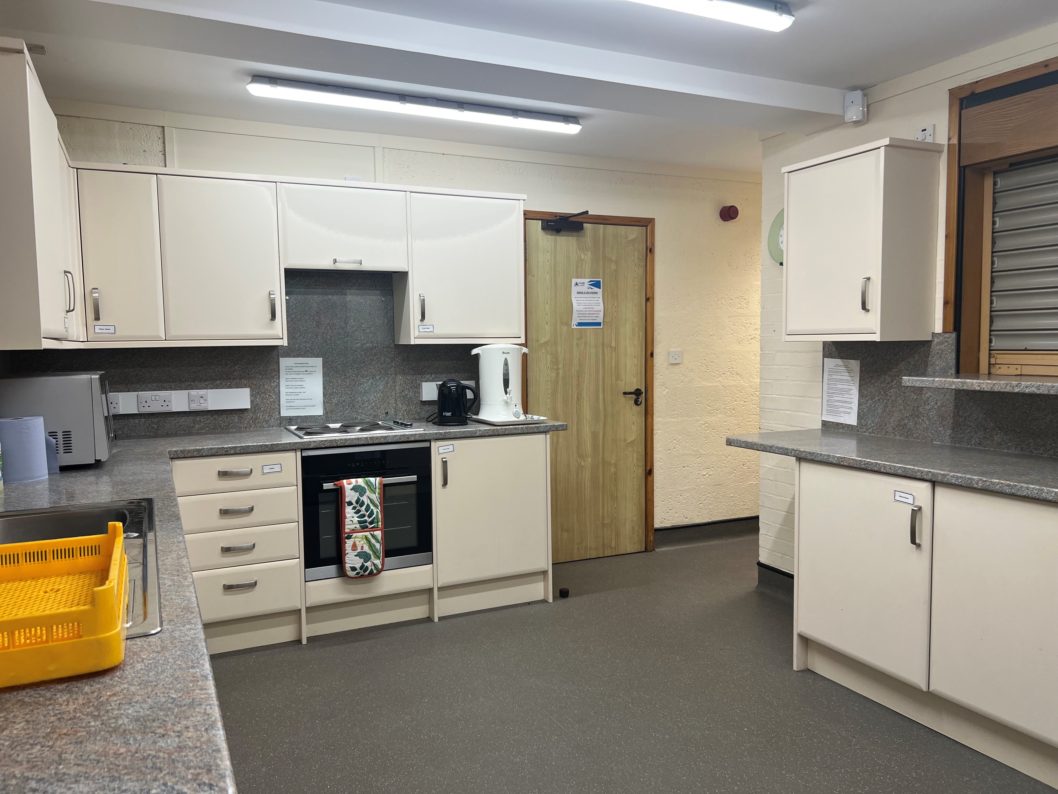 The kitchen boasts a spacious layout, offering ample countertop space and a multitude of cupboards. It is equipped with essential appliances, including a sink, an oven with a hob, a microwave, and a kettle. Additionally, there is a serving hatch that connects to the Ashton room/main hall.