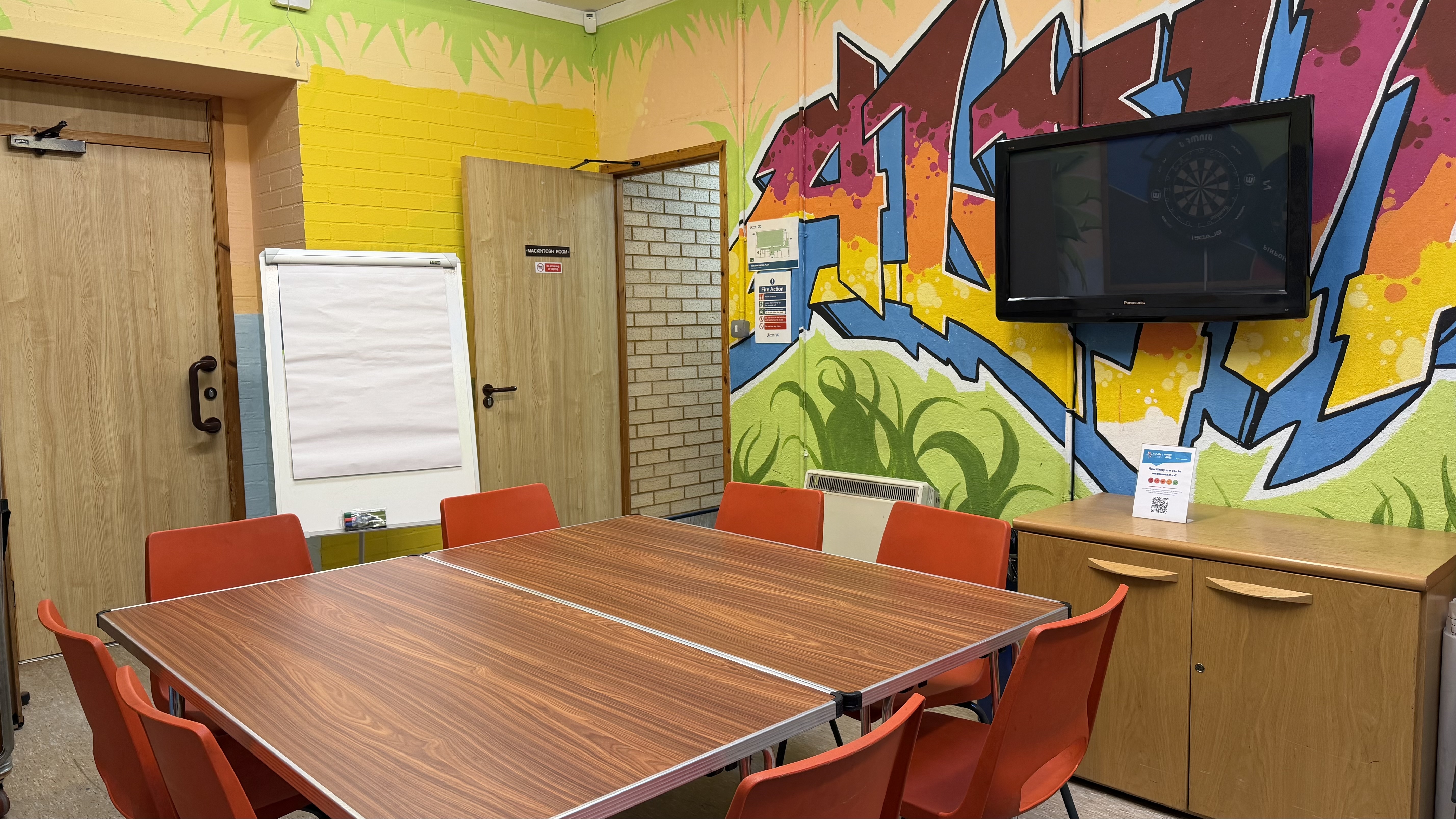 Square table in middle of room with eight orange chairs around. Wall-mounted TV to the wall to the left with flipchart next to door. Room decorated with colourful mural in graffiti style.