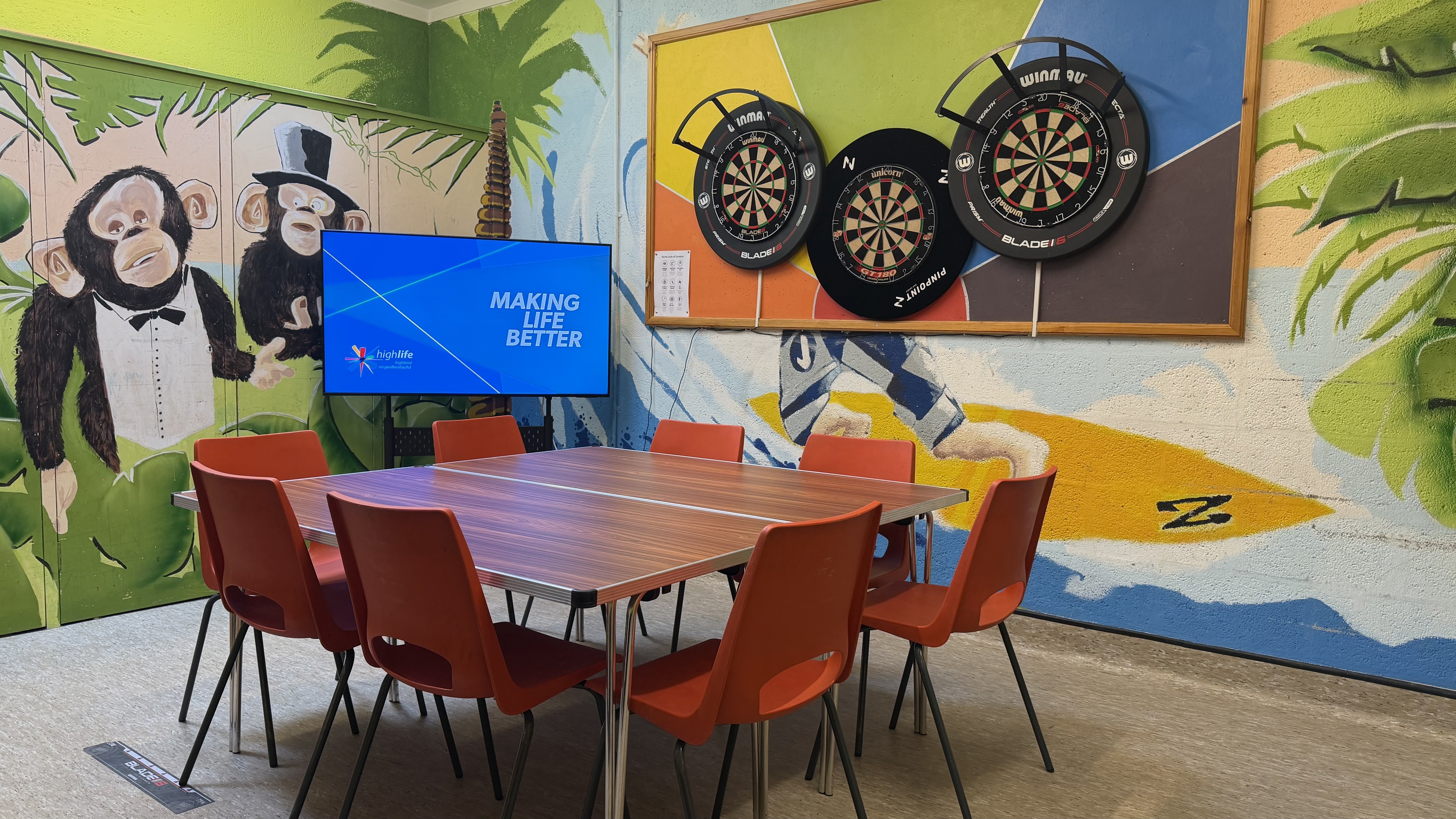 A brightly decorated room with a table and red chairs in the centre. A large screen shows the text “Making Life Better,” and three dartboards hang on a colourful wall featuring painted animals and palm trees.