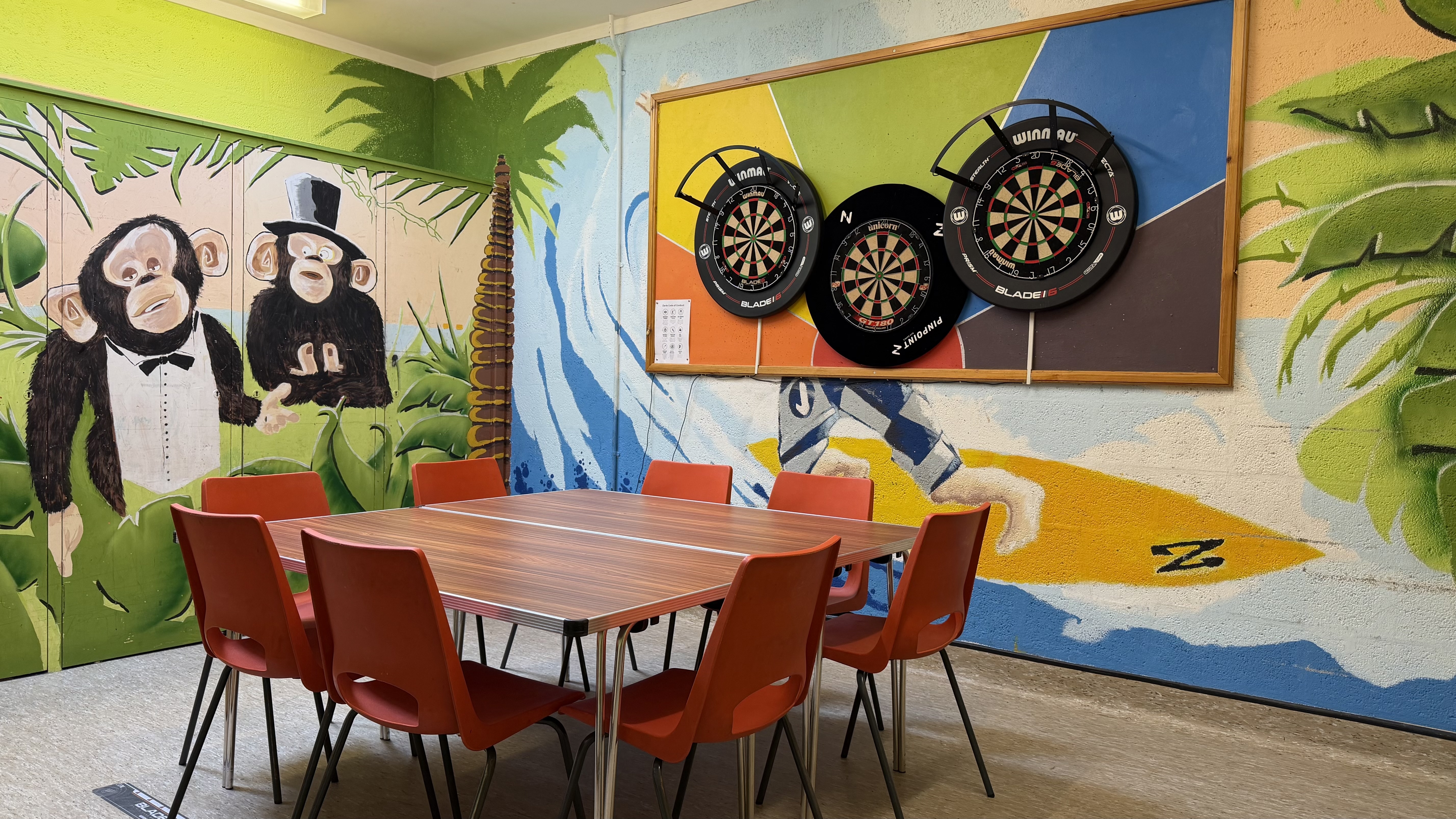 Eight chairs around square table in middle of room. Room is colourful with jungle mural to the left and three dart boards are mounted on the back wall. Small room for drop-in use.