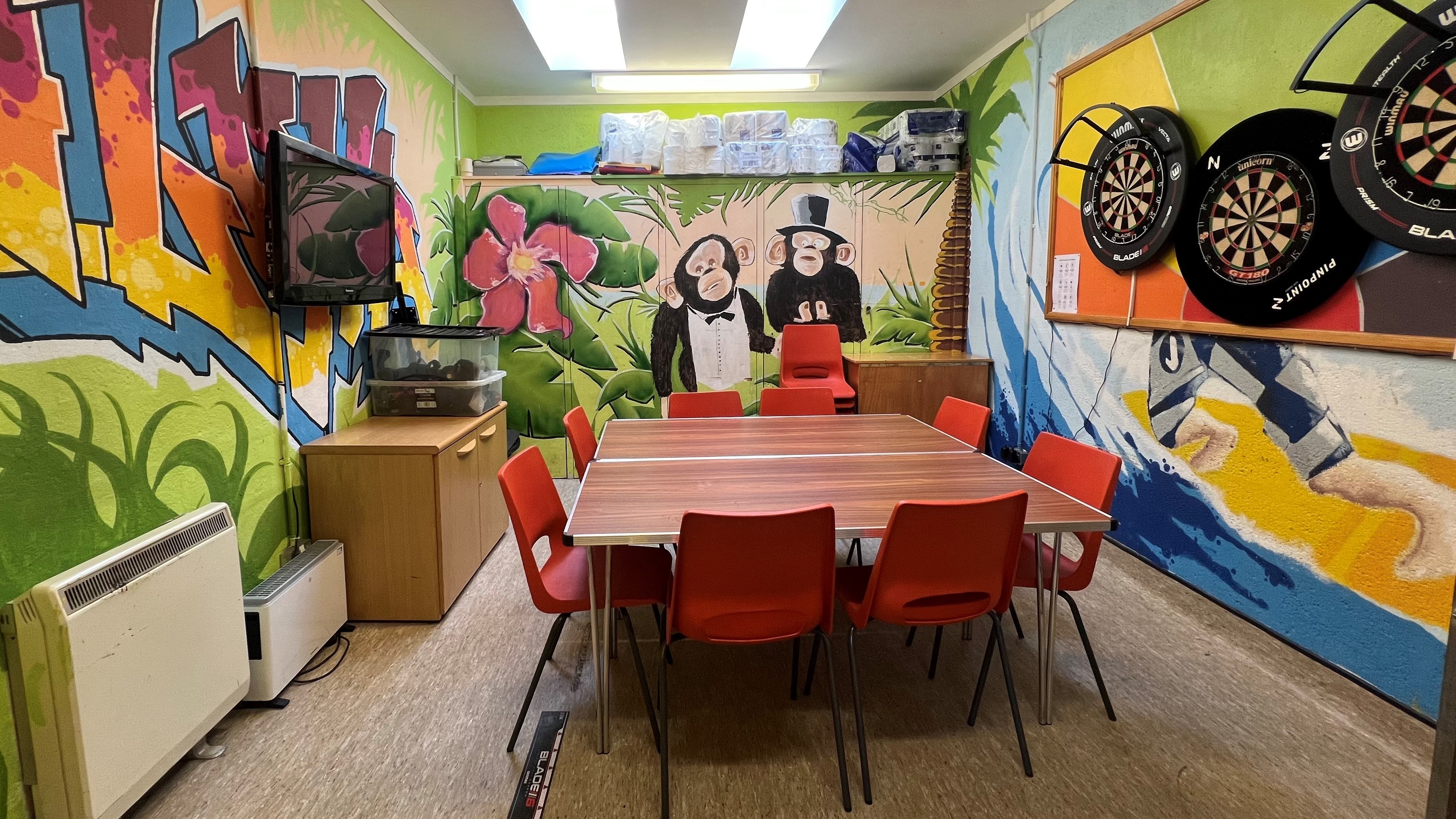 The MacKintosh room is an inviting space adorned with vibrant paintings on the walls and featuring three dartboards along one side. Centrally located are large tables surrounded by chairs, complemented by two low-level cupboards positioned against the walls and in the corner.