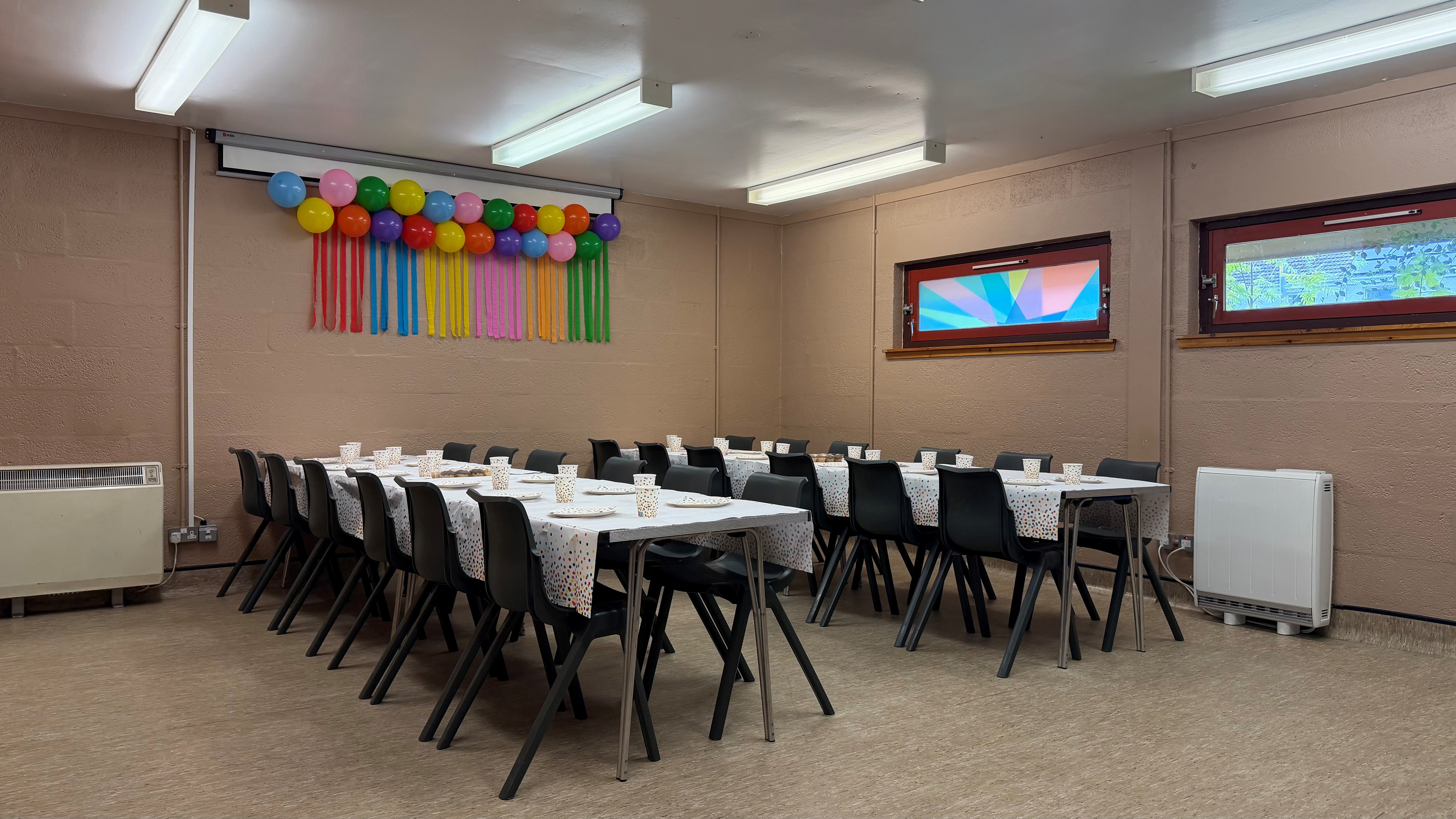 Tables set for a children’s party, with colourful party balloons hanging on wall, paper plates and cups at each place.