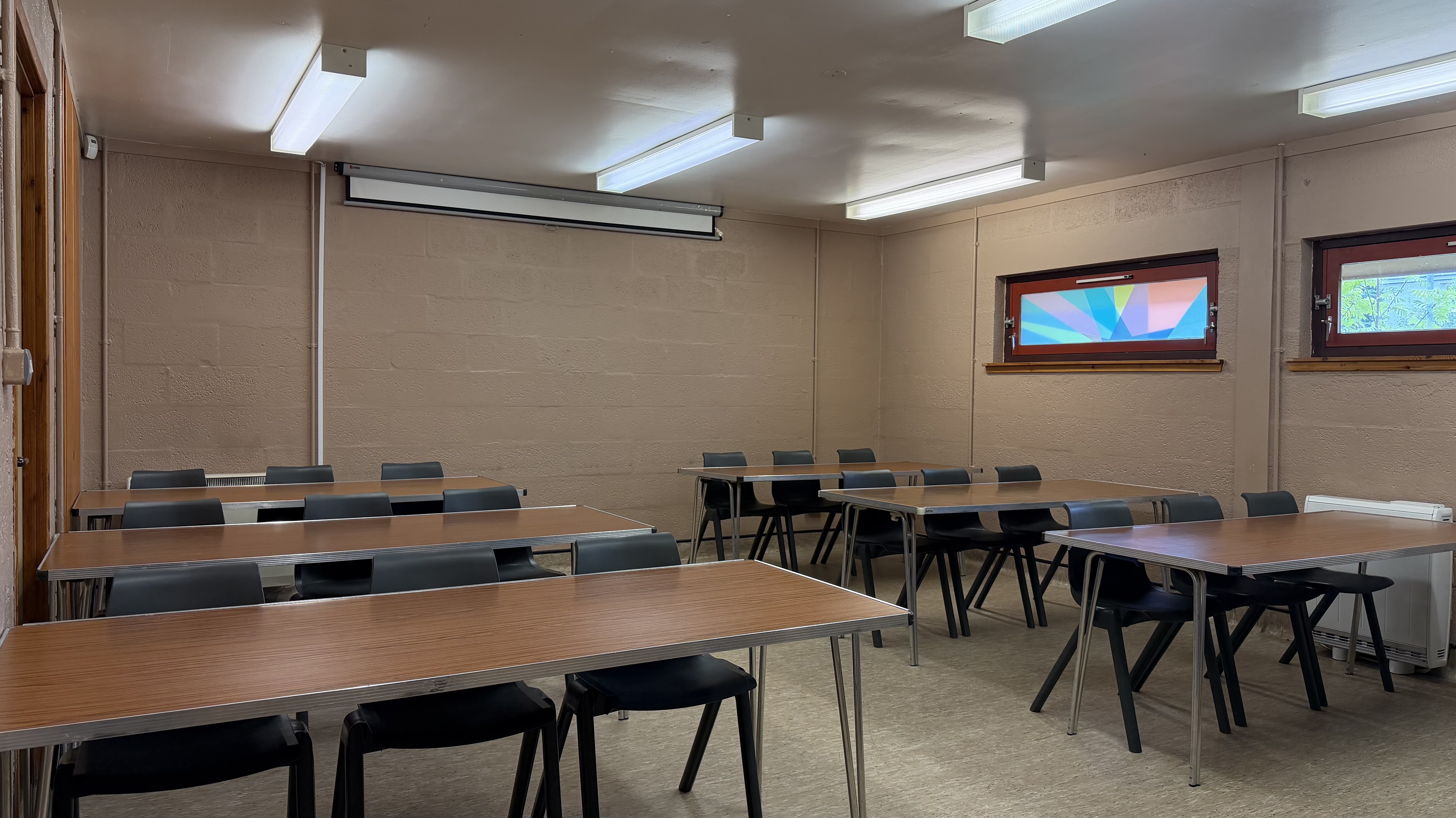 Classroom style set up in room. Six tables with three chairs under each table. two rows of three tables with gap in between.