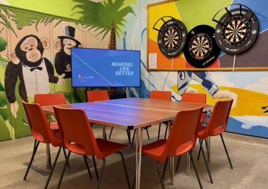 A brightly decorated room with a table and red chairs in the centre. A large screen shows the text “Making Life Better,” and three dartboards hang on a colourful wall featuring painted animals and palm trees.