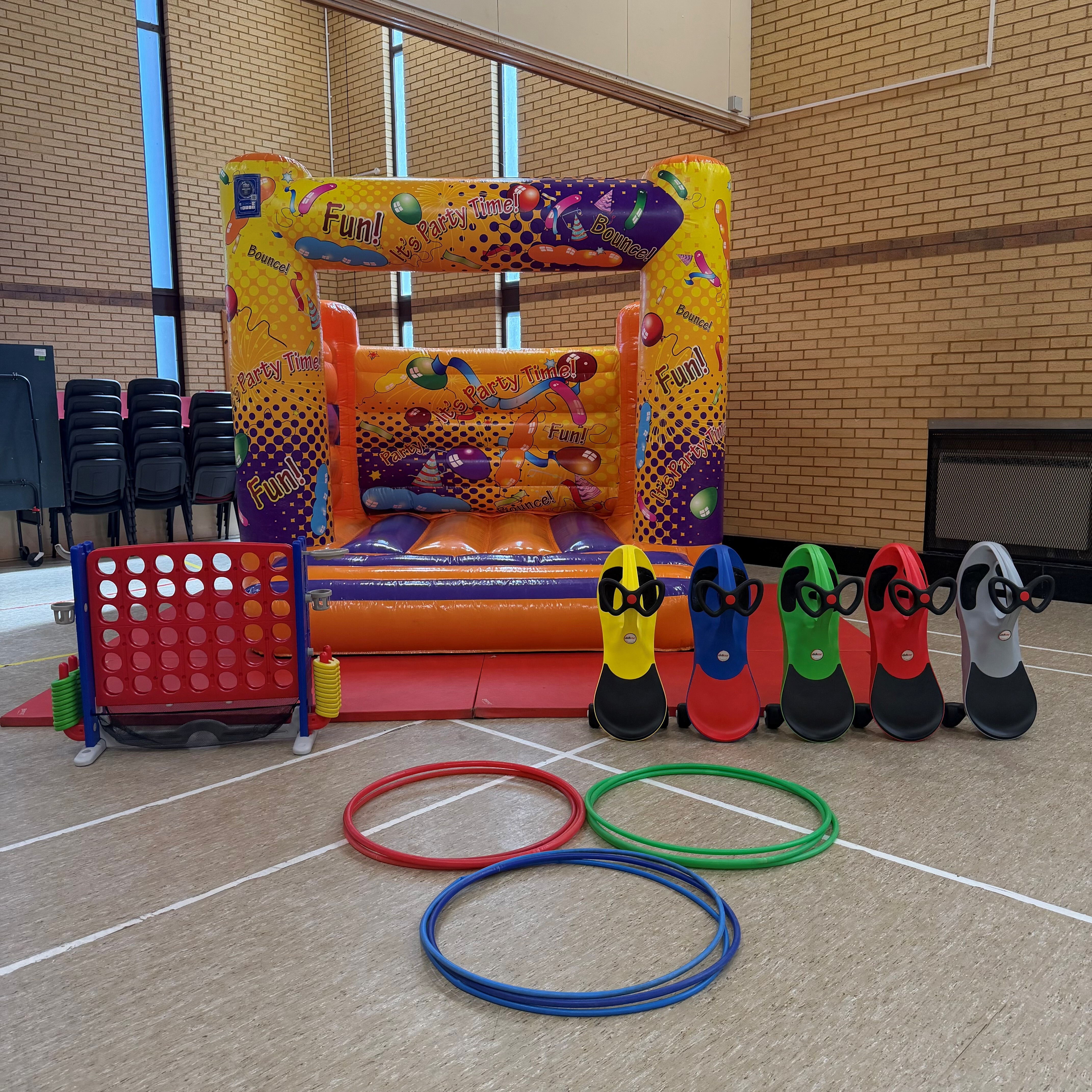 Birthday party equipment set up in a sports hall, including a bouncy castle, giant Connect 4, ride-on toys, and hula hoops.