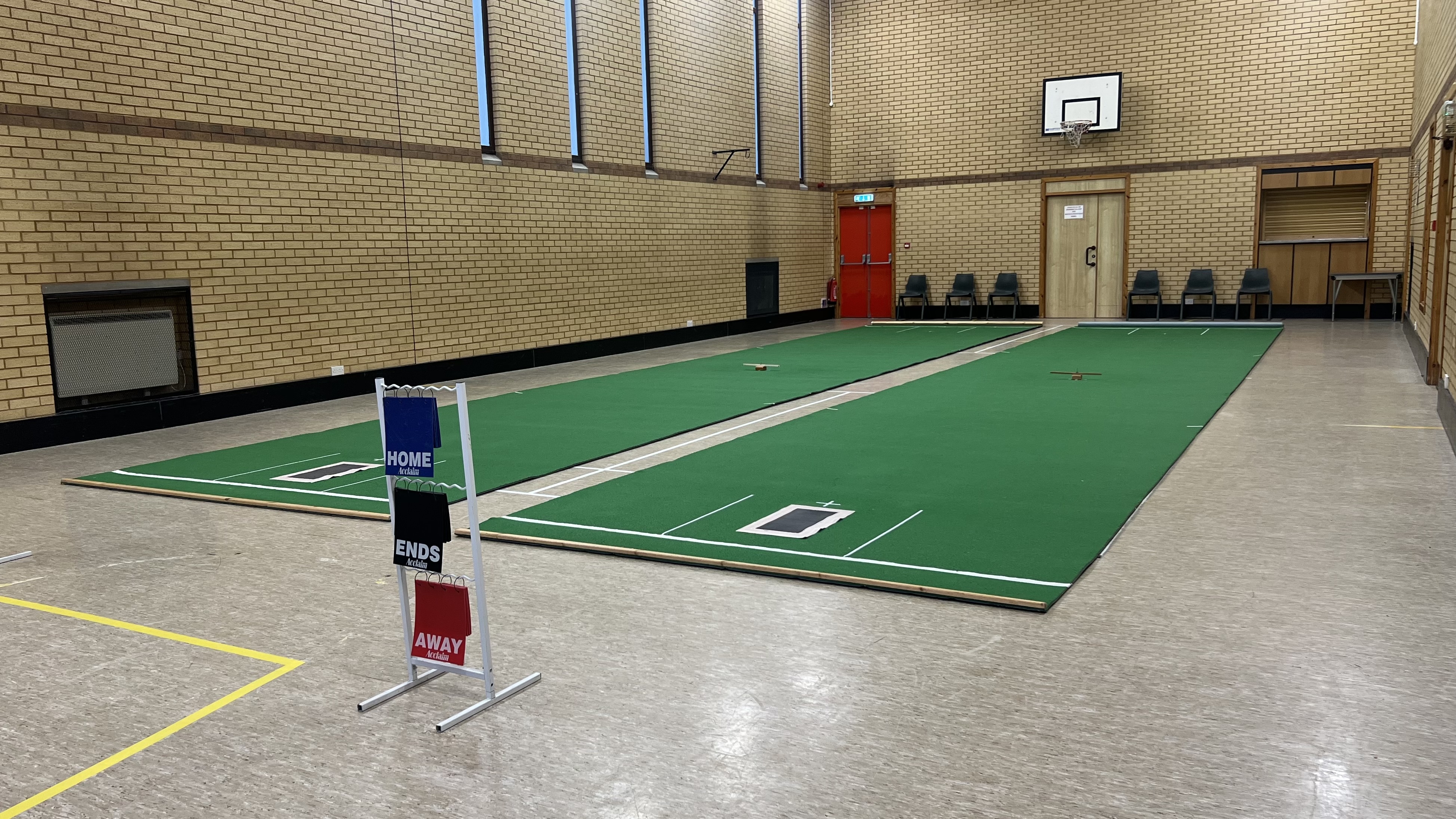 Two long indoor bowling mats laid out in a sports hall with chairs along the wall.