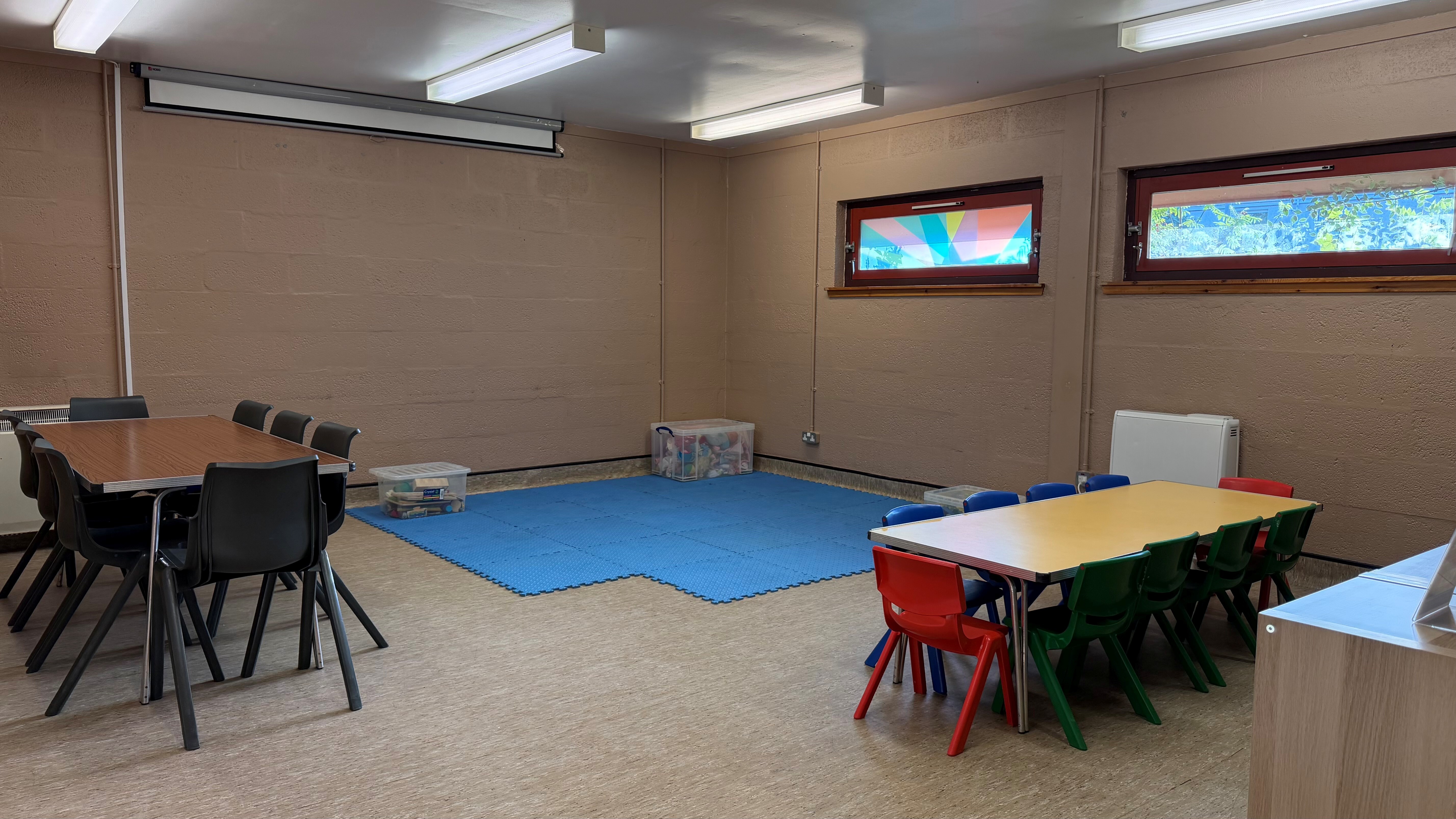 King Duncan's Room set up for a creche, with a blue foam play mat in the centre, storage boxes of toys, a low table with red and green children's chairs, and a separate table with adult chairs along the wall.
