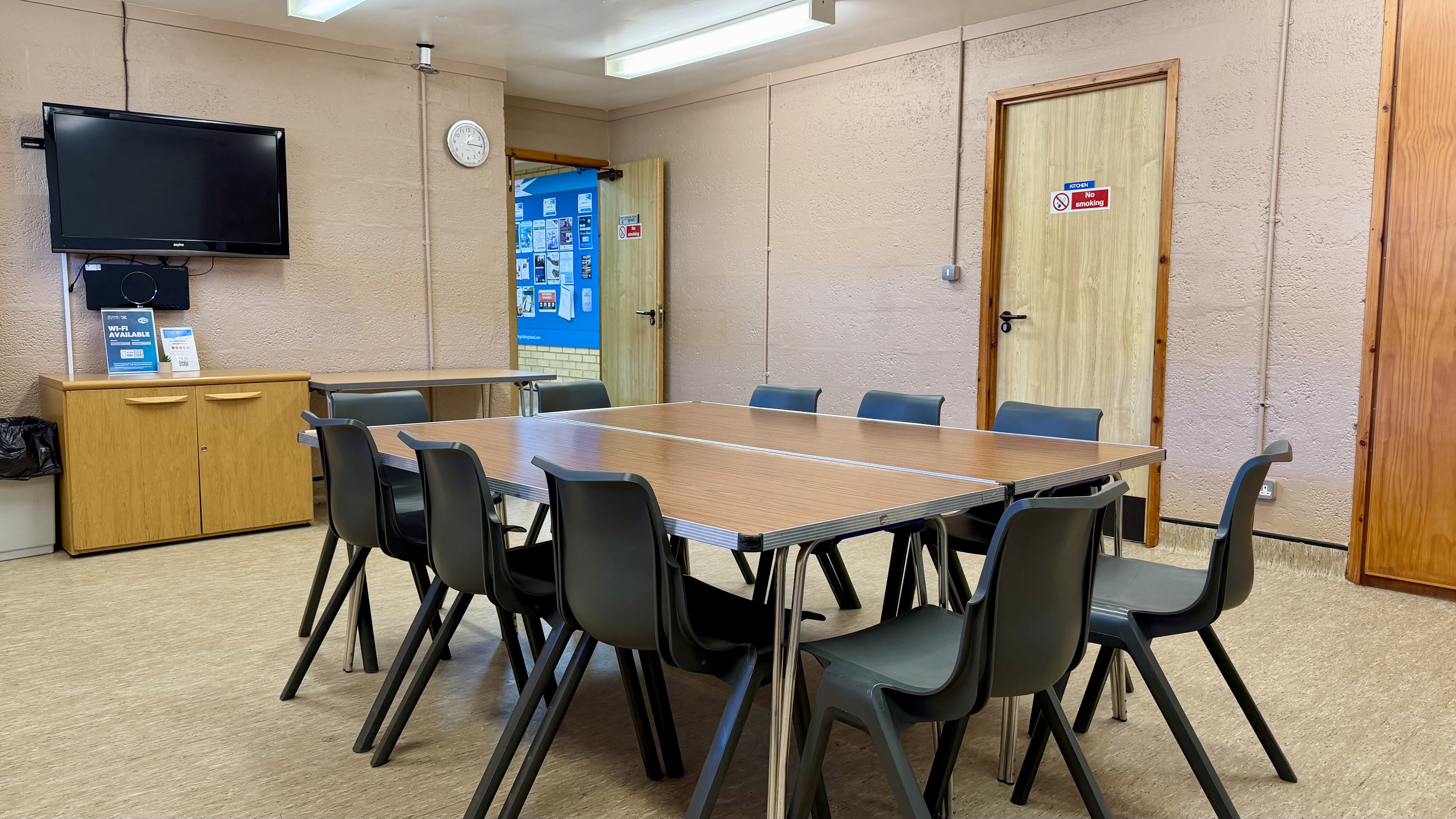 A meeting room with a large central table, chairs, a TV on the wall, and noticeboards by the door.