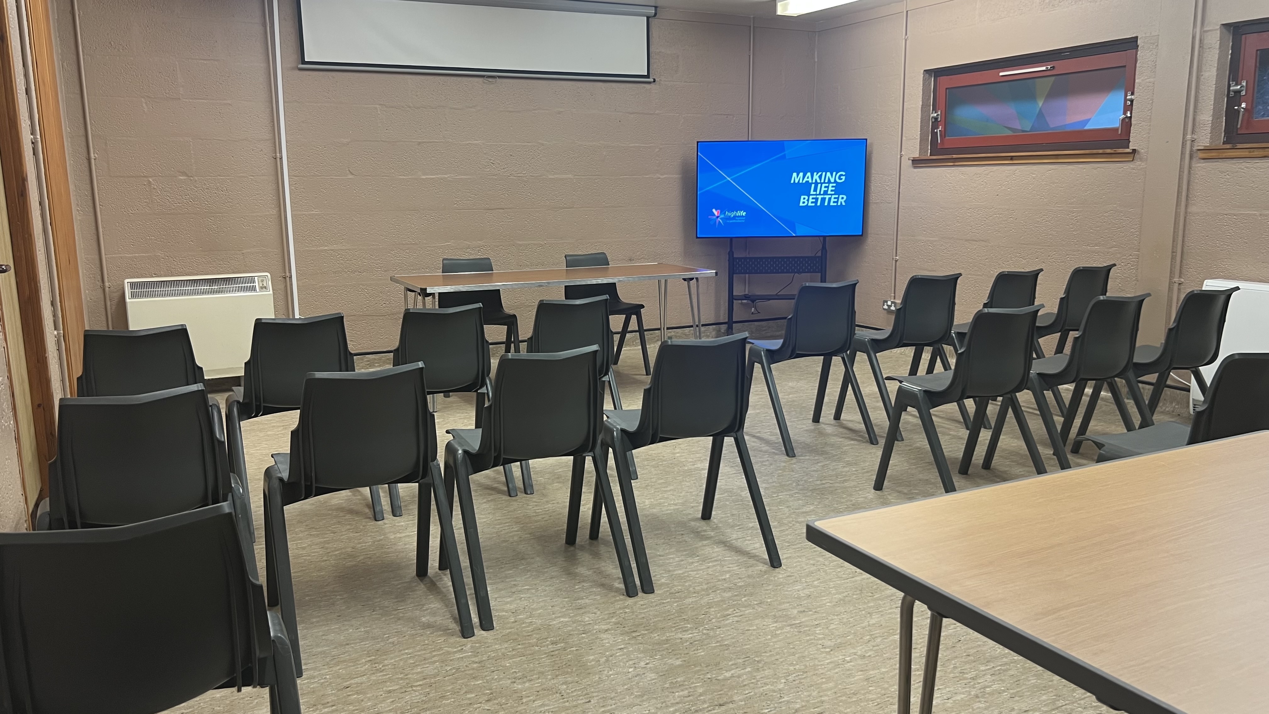 A meeting room with rows of chairs facing a table and a screen displaying “Making Life Better.”